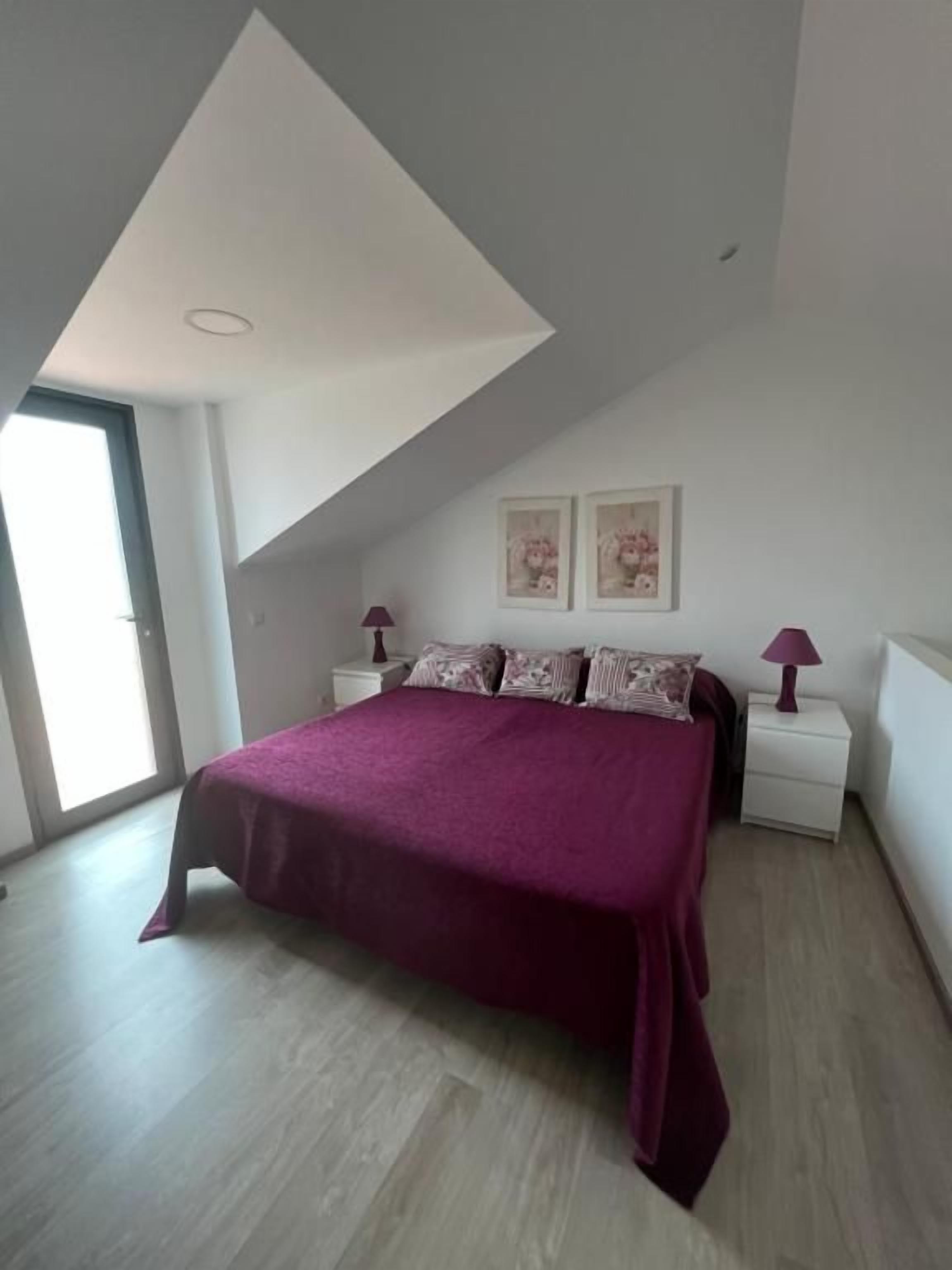 A bedroom with a sloped ceiling, a bed with a purple bedspread, two bedside lamps, and two framed pictures on the wall.