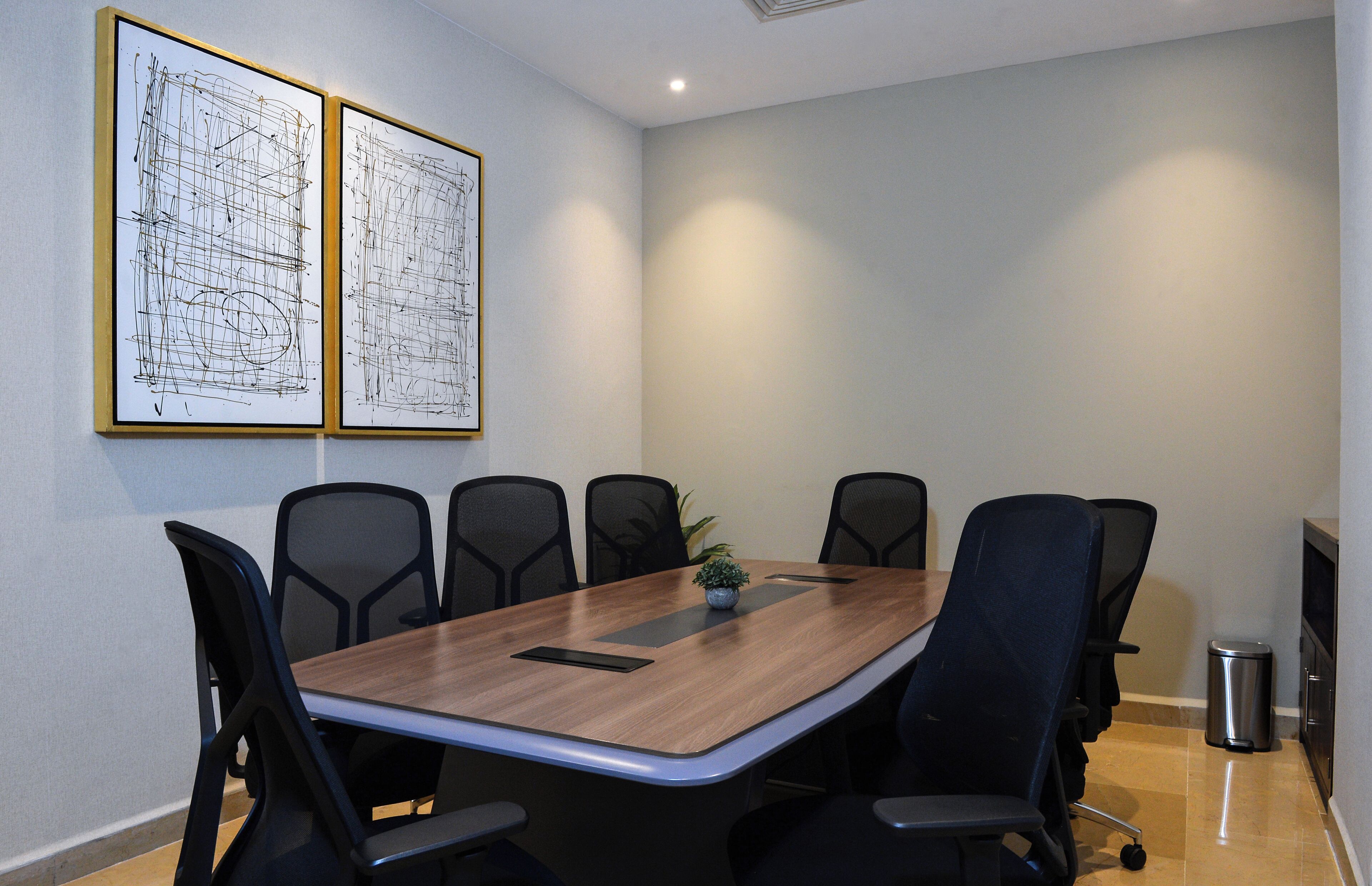 A conference room with a wooden table, six black chairs, two framed architectural drawings on the wall, and a small plant on the table.