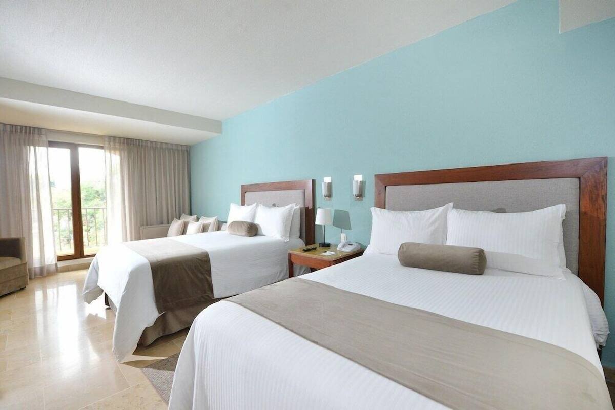 Ein Hotelzimmer mit zwei Betten, einem Schlafsofa, einem Fenster mit Vorhängen und einem Nachttisch mit Lampe.