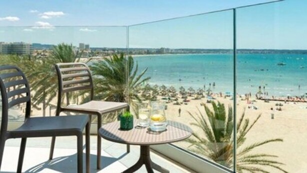 A balcony with two chairs and a table, overlooking a beach with people and boats. - Hotel Las Arenas (Palma de Mallorca)
