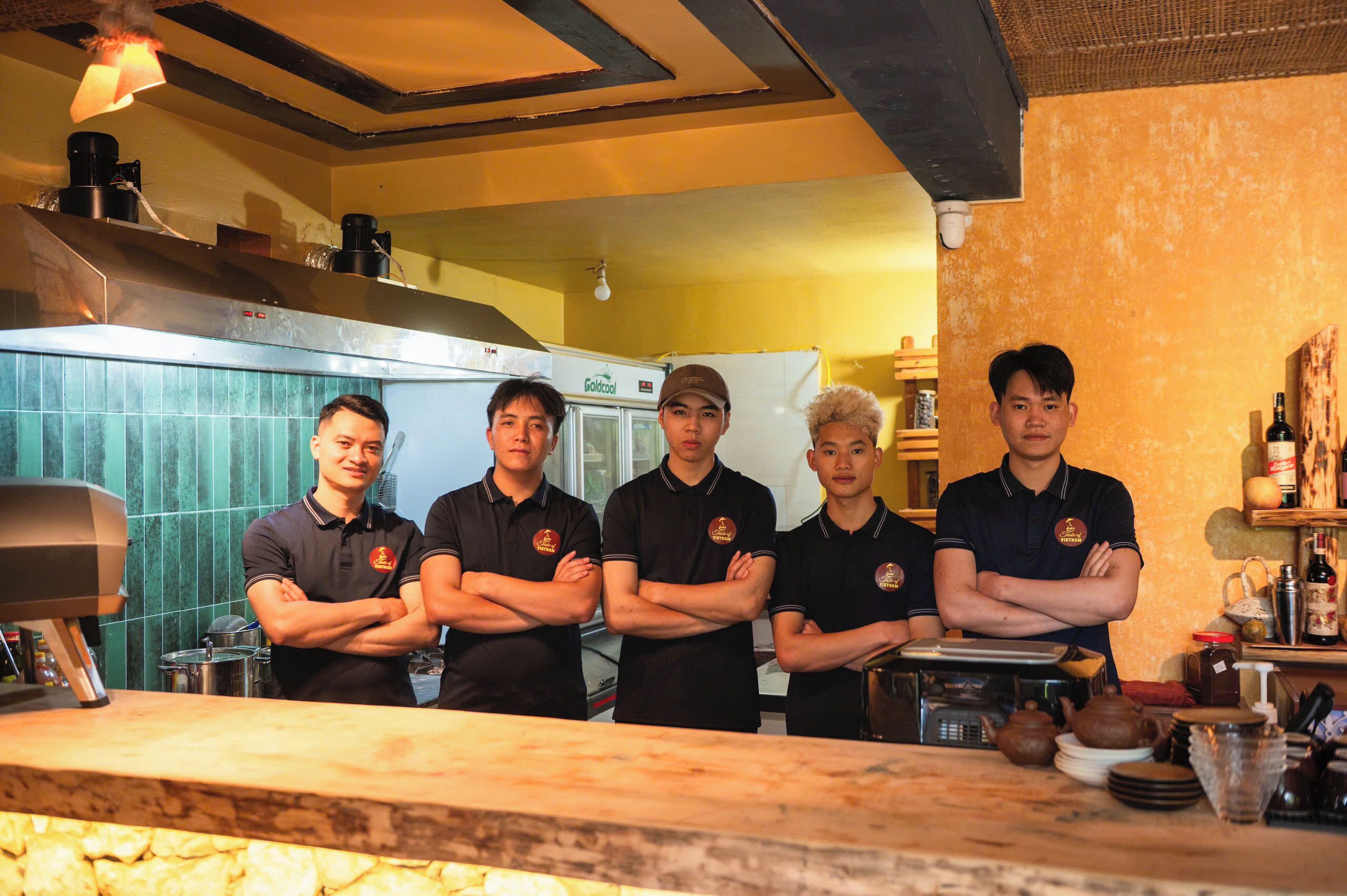 Un grupo de personas de pie en una cocina con paredes de azulejos, una encimera de madera y diversos electrodomésticos.