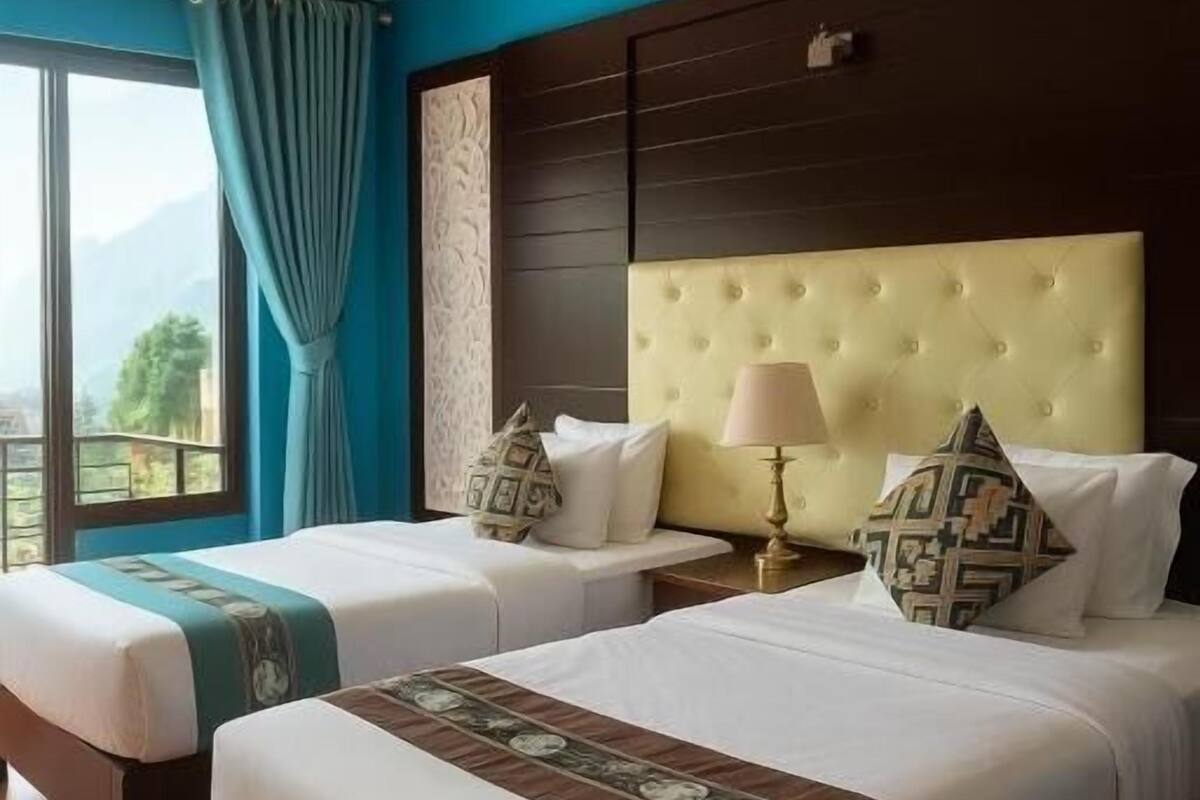A hotel room with two beds, a large window with curtains, a view of mountains, and a dark wooden headboard.