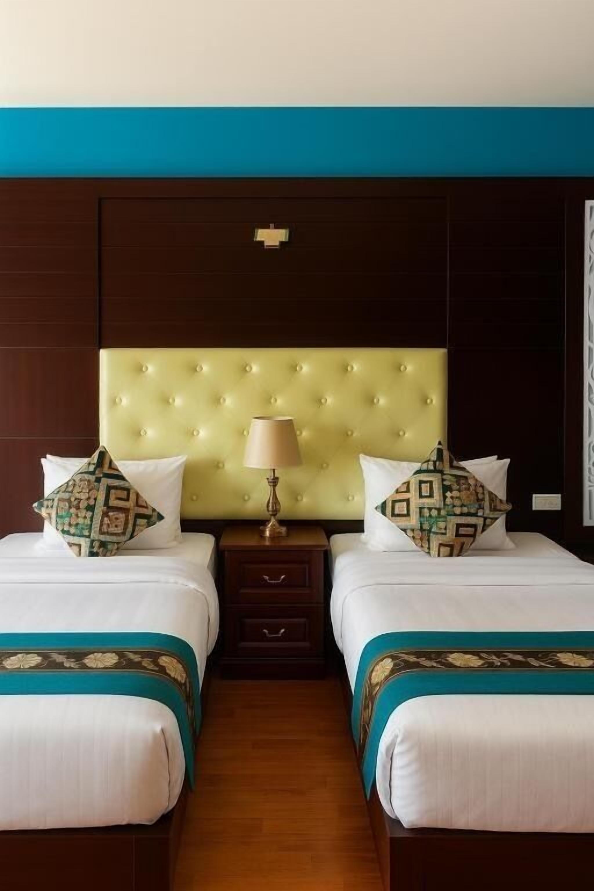 A hotel room with two beds, a tufted headboard, a nightstand with a lamp, and a decorative pillow.