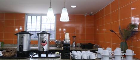Une cuisine avec des carreaux de céramique orange, deux machines à café et un assortiment de tasses et d’assiettes blanches.