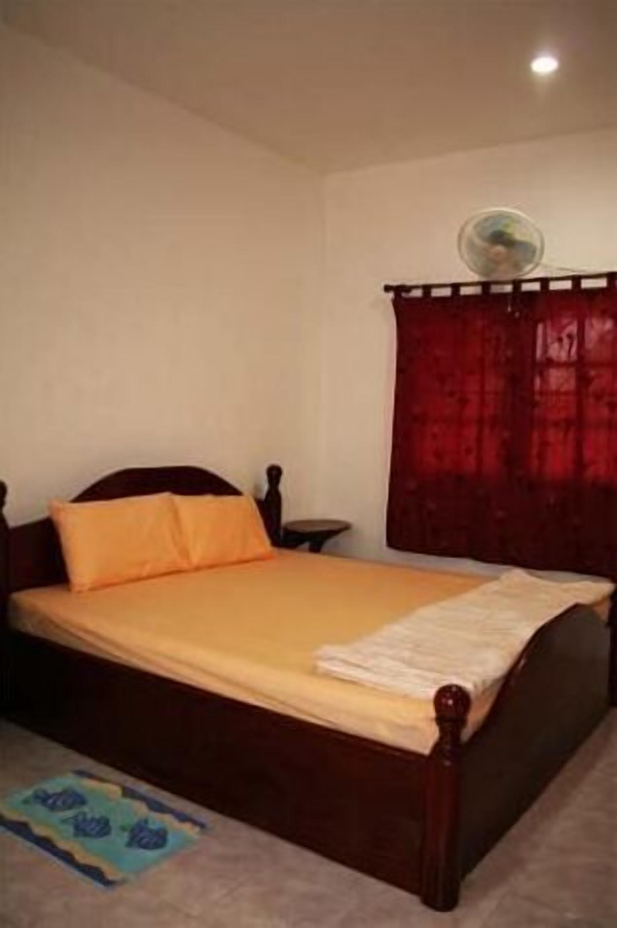 A bedroom with a wooden bed, orange bedding, a small bedside table, a fan, and red curtains.