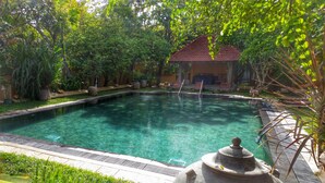 A clear swimming pool surrounded by lush greenery and a small building with a red roof. - Jetwing Ayurveda Pavilions (Negombo)
