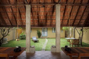 Un espacio al aire libre cubierto con techo de paja, un pequeño jardín con césped, una silla de descanso y una mesa con una toalla y un tapete enrollado.