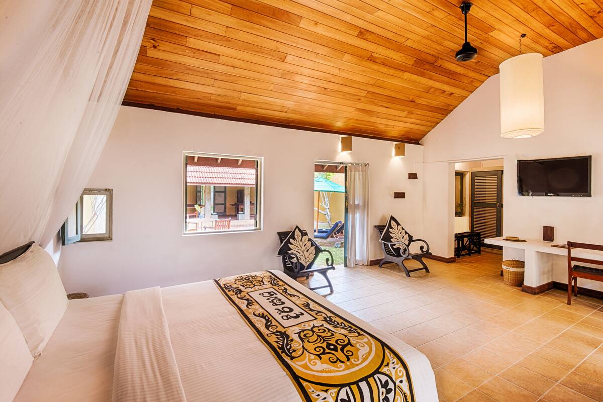 A bedroom with a large bed, wooden ceiling, two windows, a TV, and a desk.