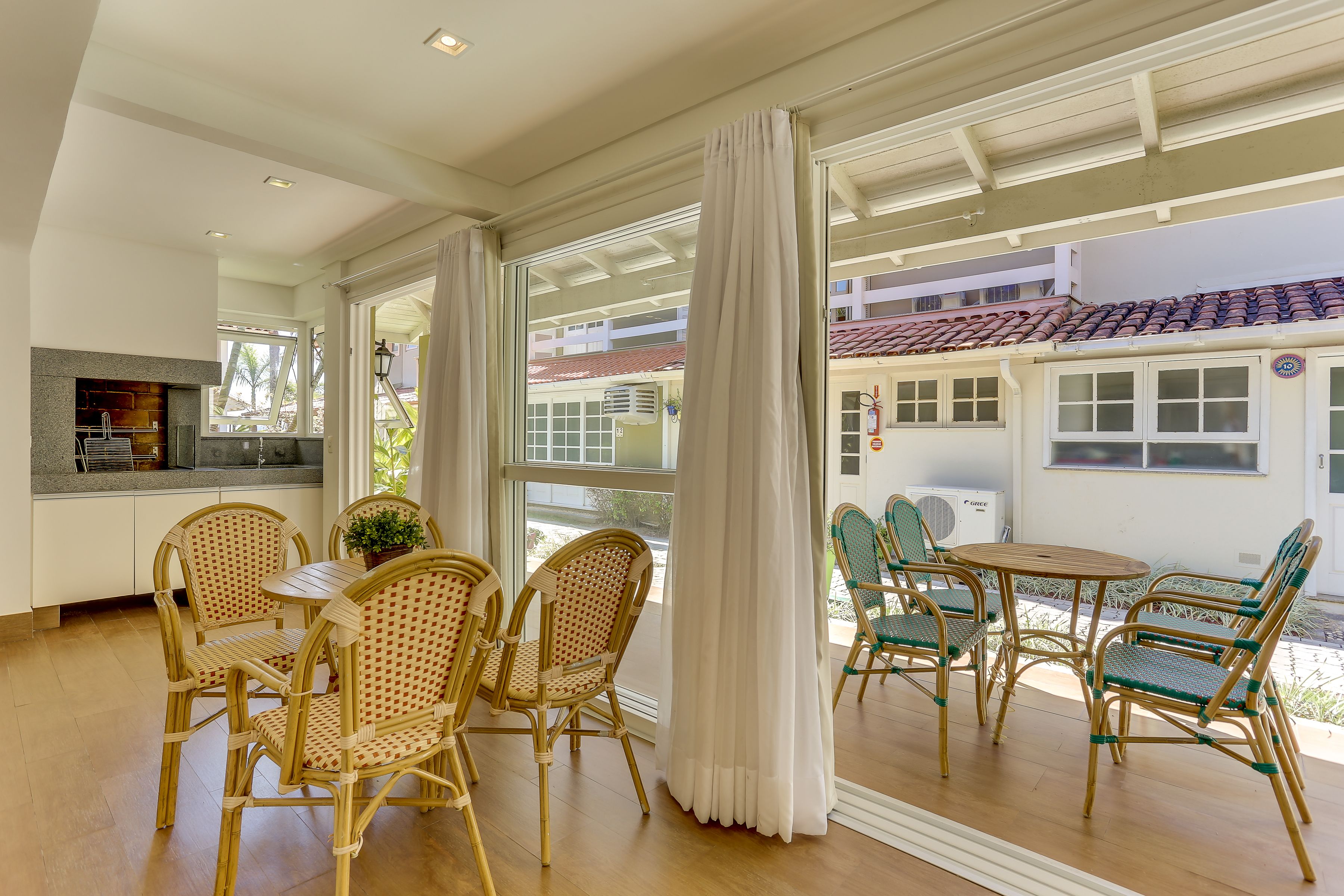 A spacious dining area with wicker chairs and a round table, large windows with white curtains, and a view of a building with a red-tiled roof.