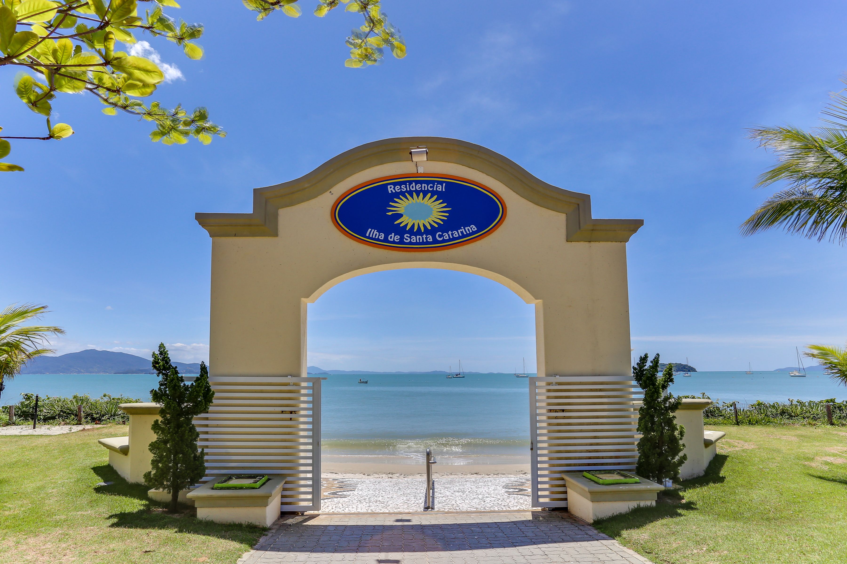 Un arco de entrada con un letrero que dice 'Residential Ilha de Santa Catarina' y vistas al mar con barcos.