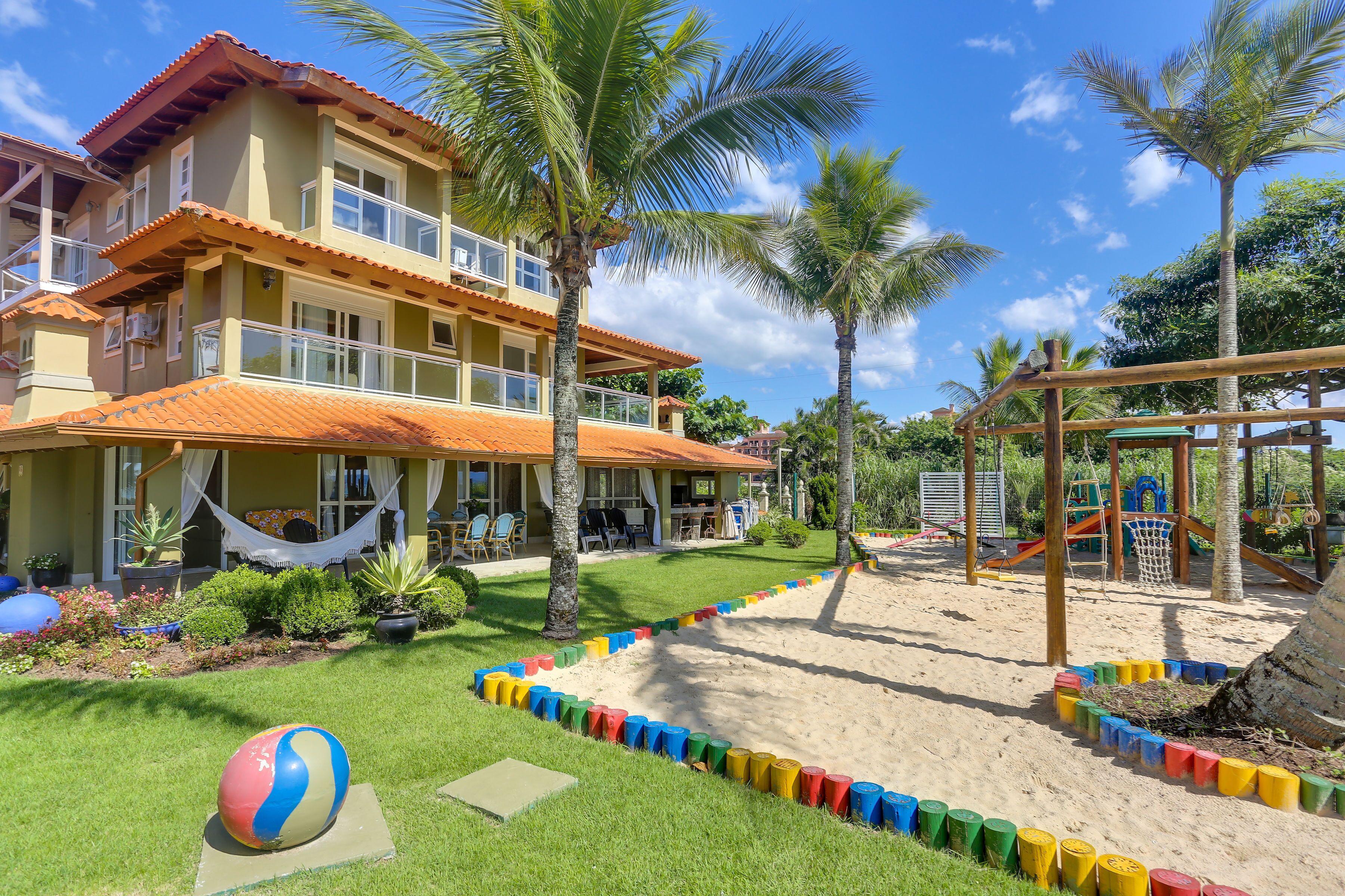A two-story building with a balcony, surrounded by palm trees and a sandy area with colorful barriers.