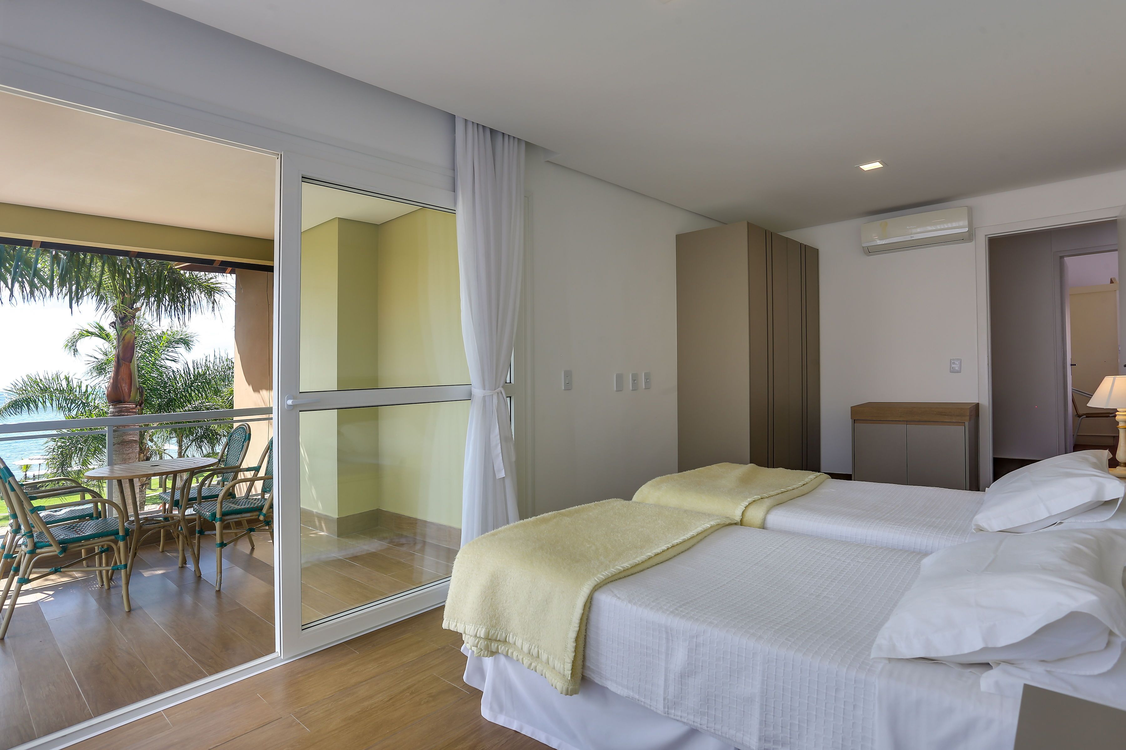 Habitación de hotel con balcón, cama con ropa blanca, mesita de noche de madera y vista a palmeras.