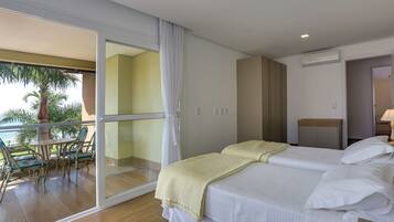 Habitación de hotel con balcón, cama con ropa blanca, mesita de noche de madera y vista a palmeras.