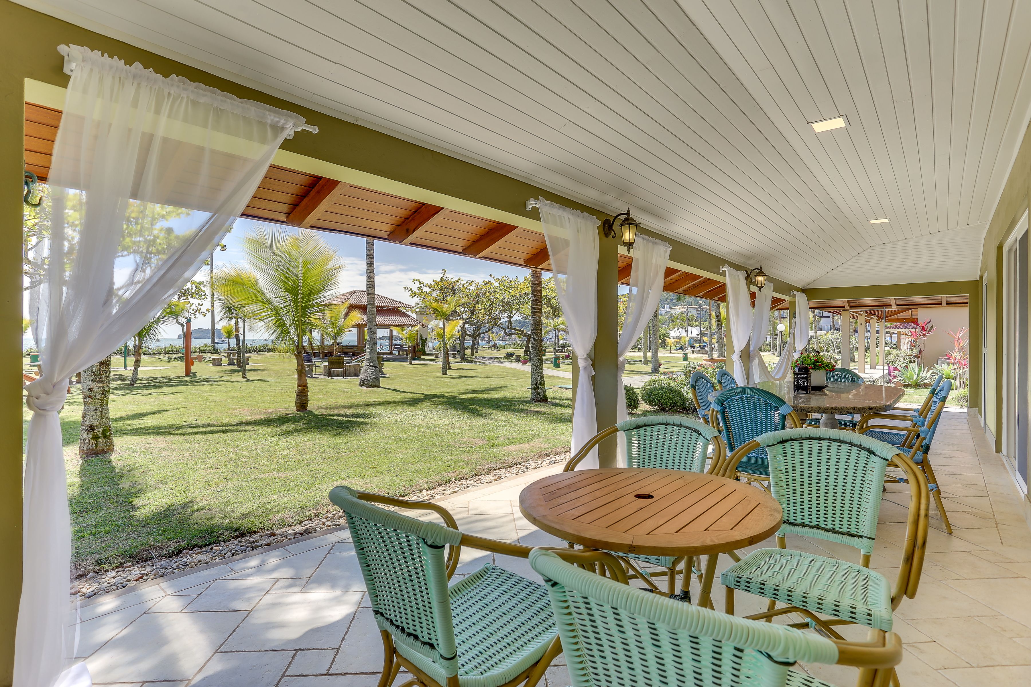 A covered patio area with wicker chairs and a wooden table, overlooking a well-maintained lawn and distant buildings.
