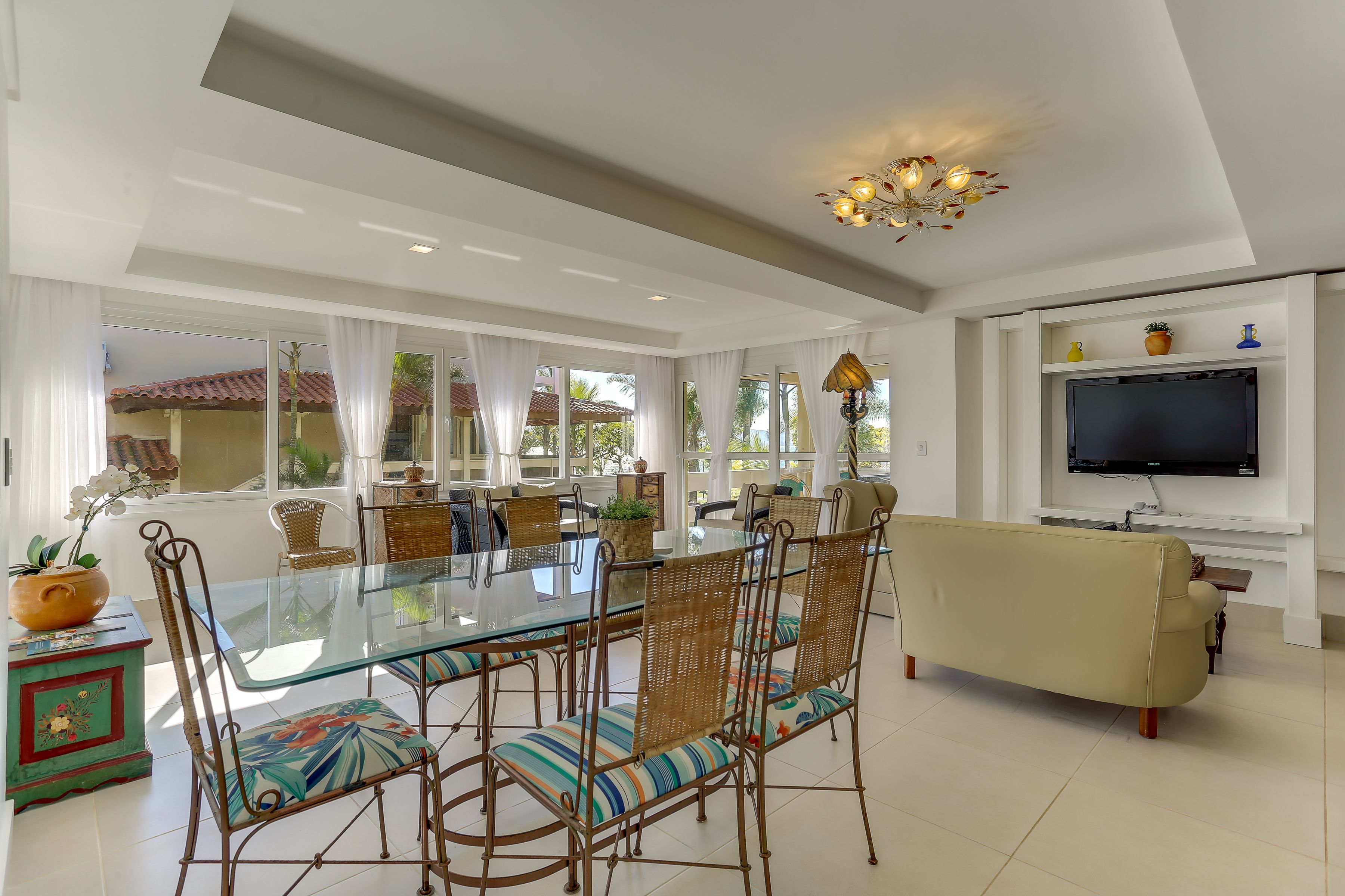 A spacious living room with a glass dining table, wicker chairs, a beige sofa, and a TV mounted on the wall.