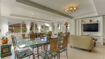 A spacious living room with a glass dining table, wicker chairs, a beige sofa, and a TV mounted on the wall.