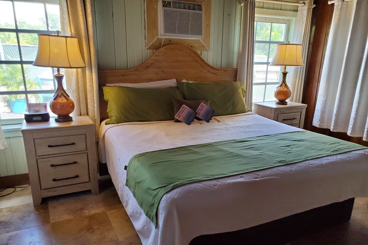 A neatly made bed with a green bedspread, two pillows, and a wooden headboard. There are two bedside tables with lamps and a small drawer.