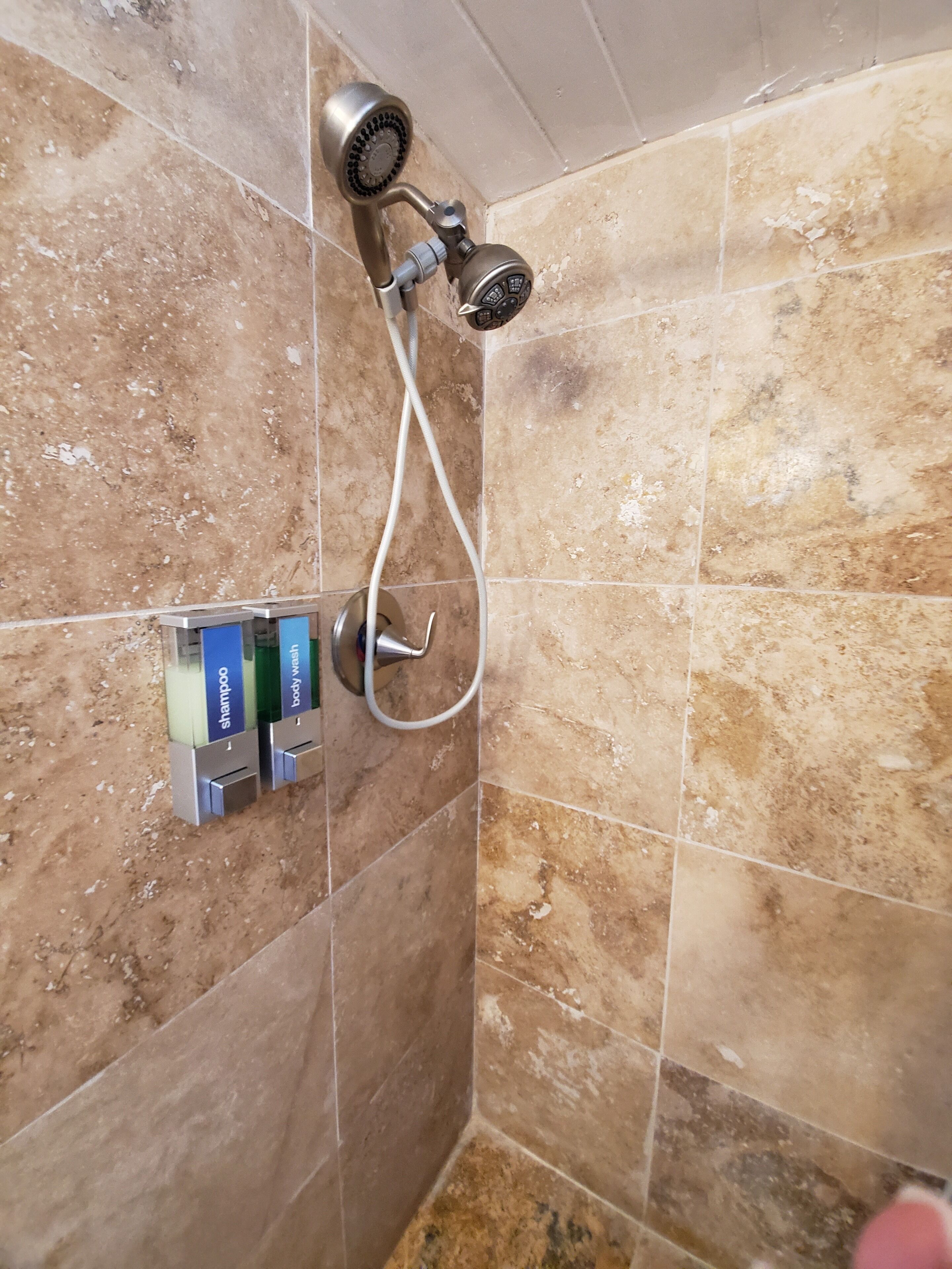 Un espace douche équipé d’un pommeau de douche à main et d’un panneau de commande fixé au mur, avec des distributeurs étiquetés pour le shampoing et le gel douche.