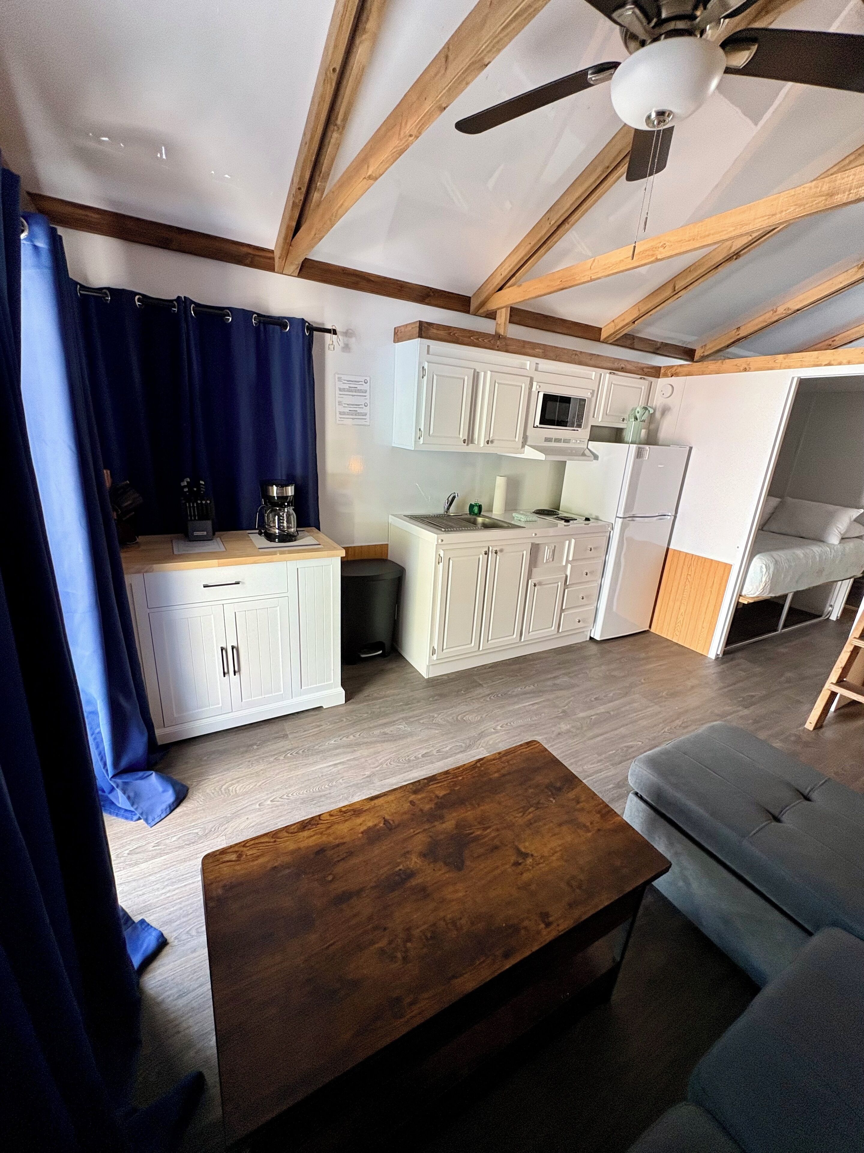 A compact kitchen with white cabinets, a microwave, and a refrigerator. There is a wooden ceiling fan, a blue curtain, and a small wooden table.