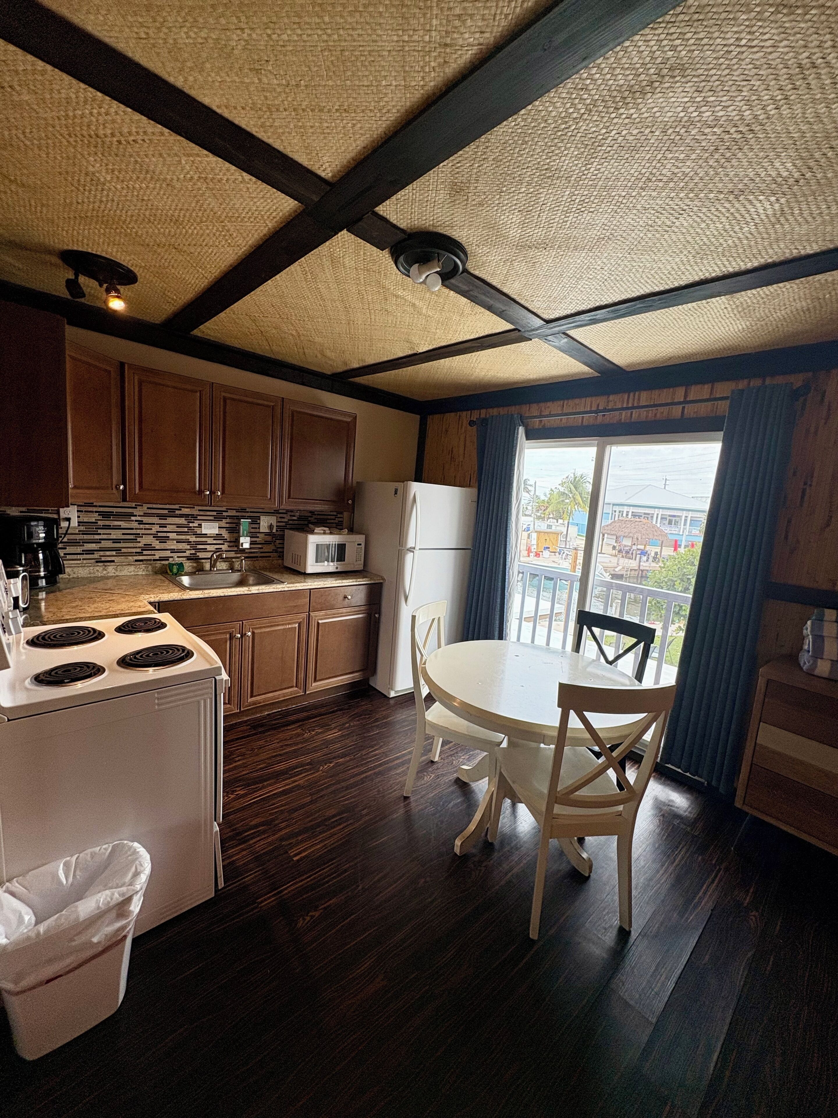 Une cuisine compacte avec des meubles en bois, une cuisinière blanche, un four à micro-ondes et un réfrigérateur. On y trouve une table à manger avec des chaises, et une grande fenêtre offre une vue sur l’extérieur.
