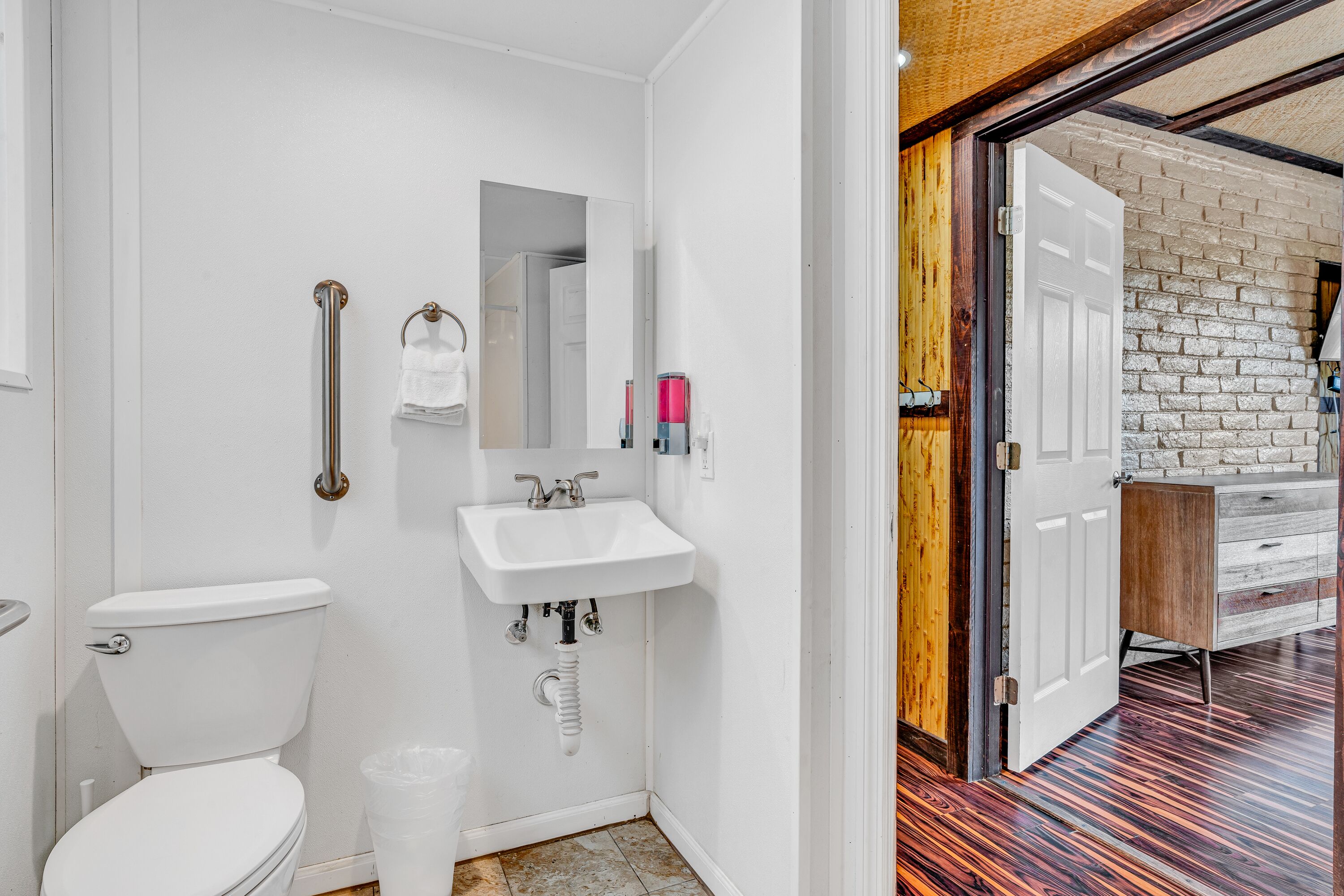Un baño con inodoro blanco, lavamanos y espejo. Una puerta de madera da a otra habitación.
