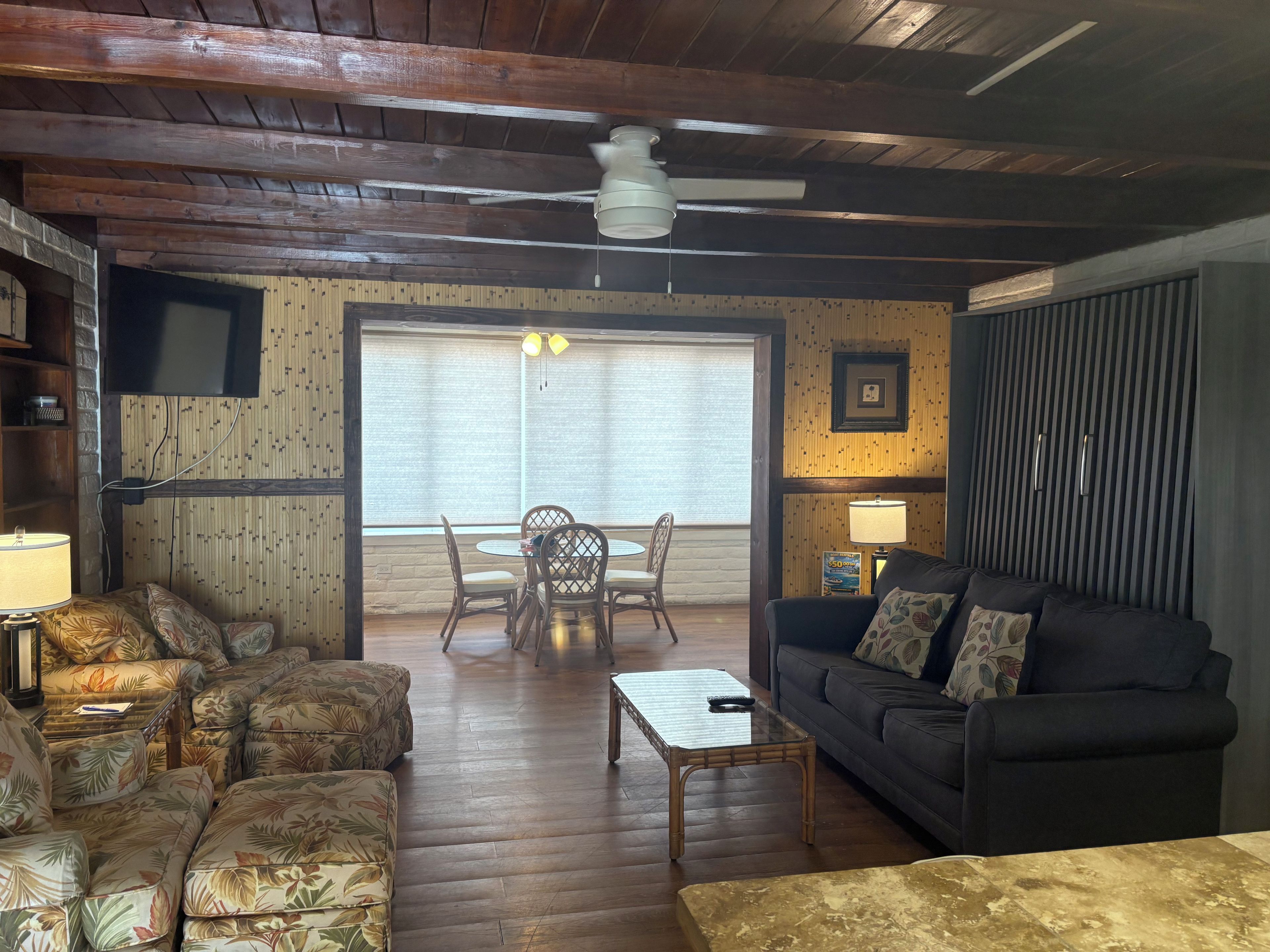 A cozy living room with a wooden ceiling, a ceiling fan, a television mounted on the wall, a dining area with a table and chairs, and a sofa with cushions.