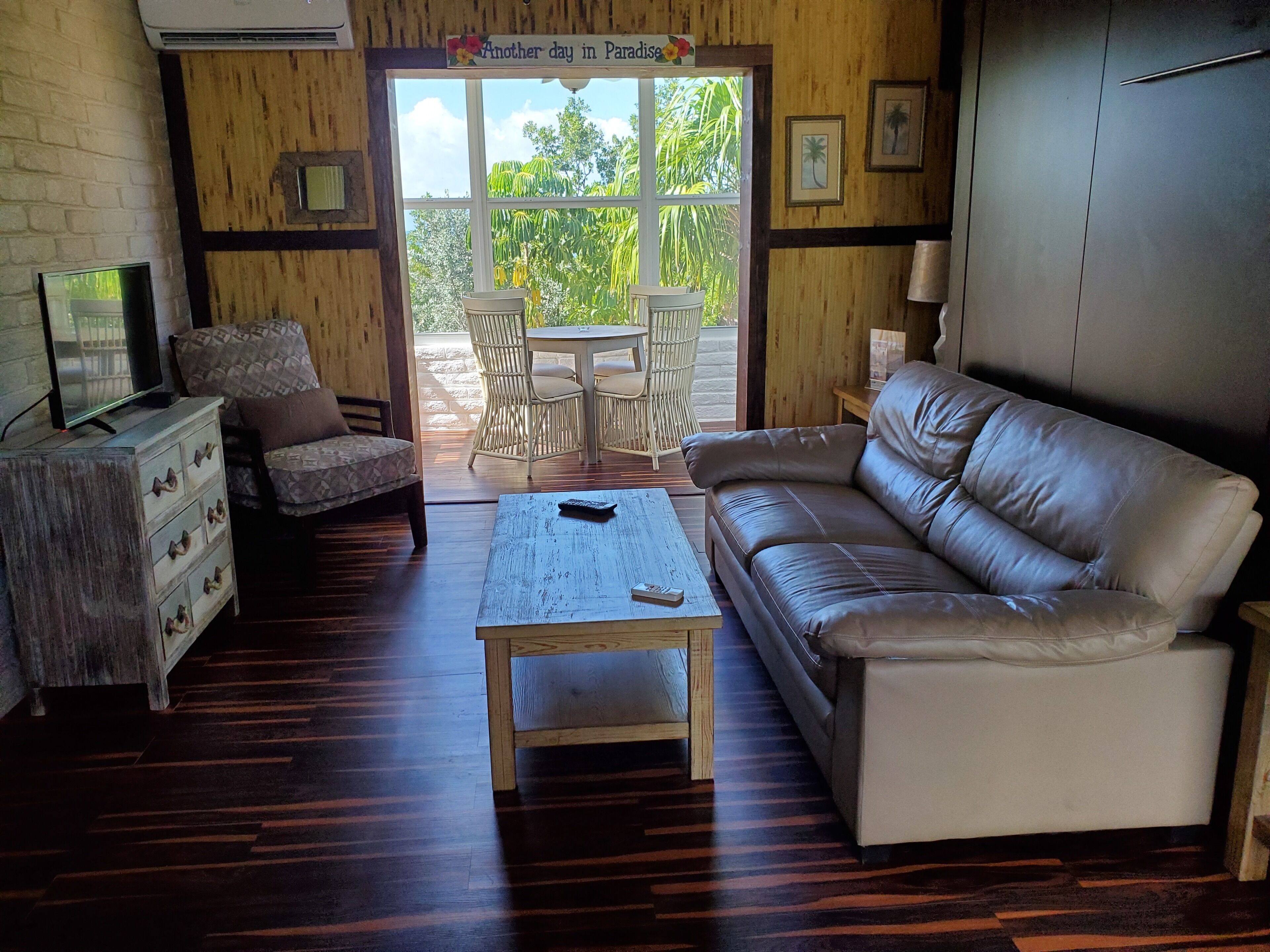 Uma sala de estar aconchegante com sofá, mesa de centro, TV sobre um armário de madeira, cadeiras de vime e vista para o exterior através de uma porta de vidro.