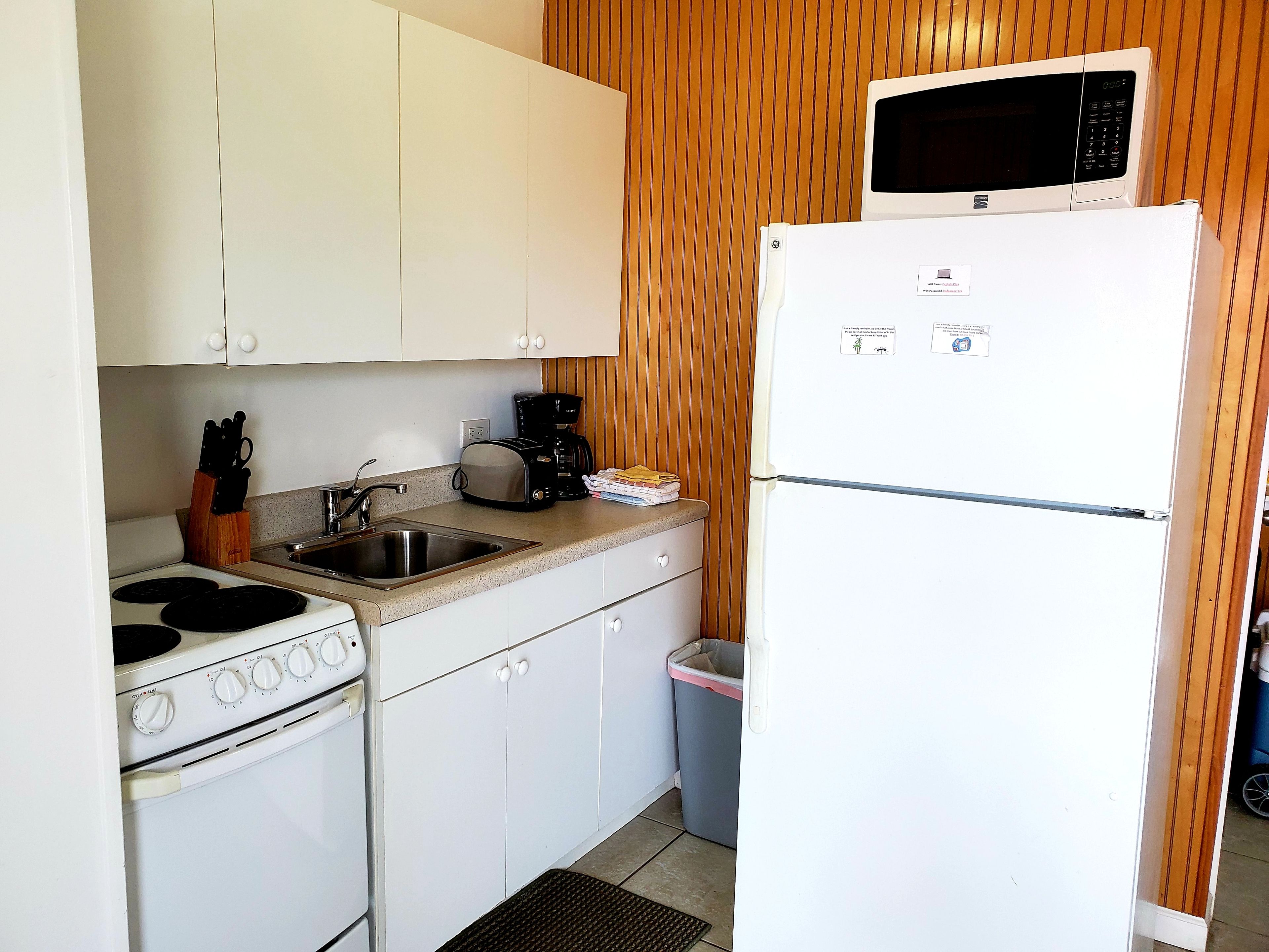 A compact kitchen with white cabinets, a stove, a sink, a microwave, and a trash bin.