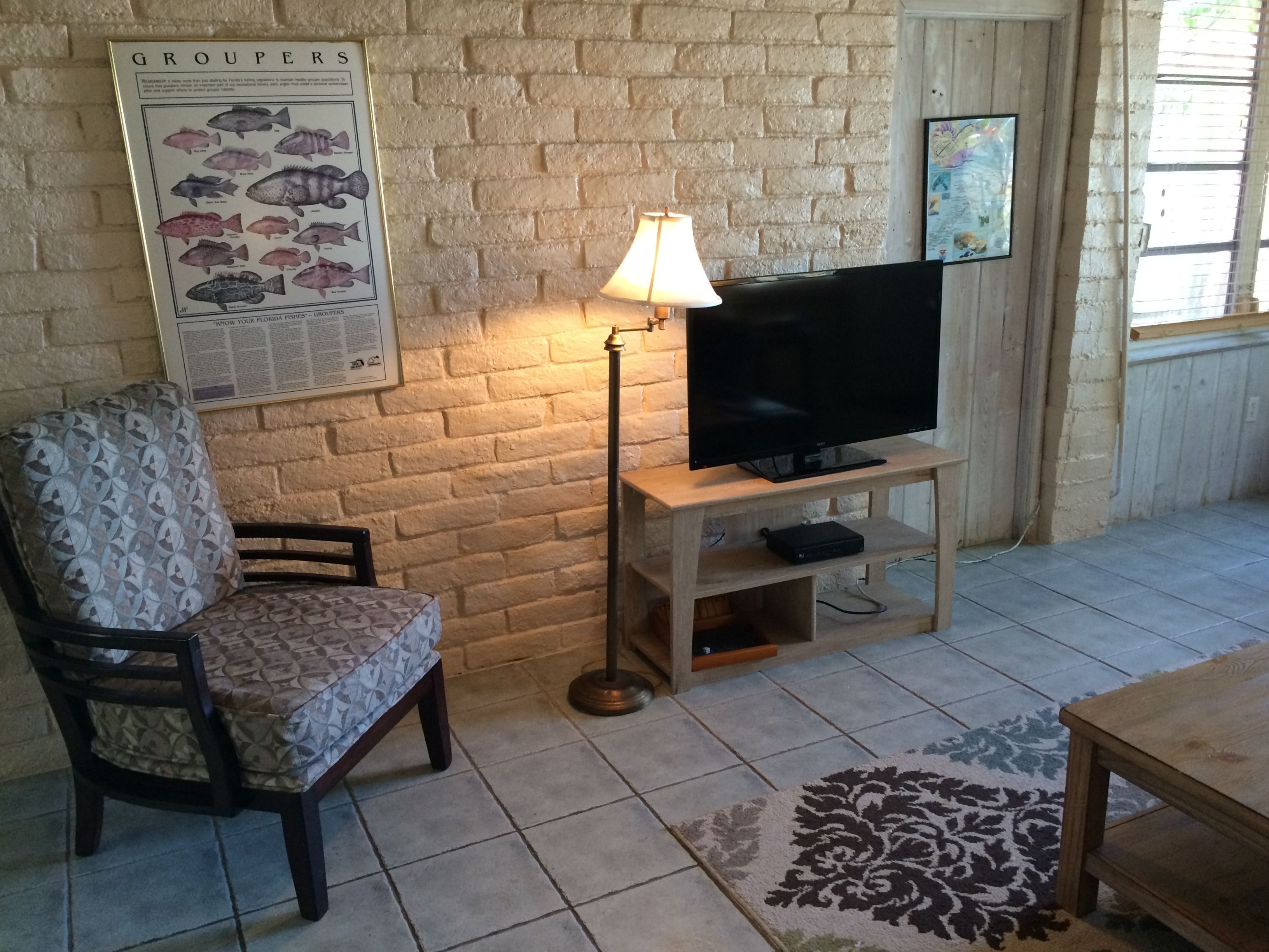 A living room with a brick wall, a television on a wooden stand, a patterned armchair, and a lamp.
