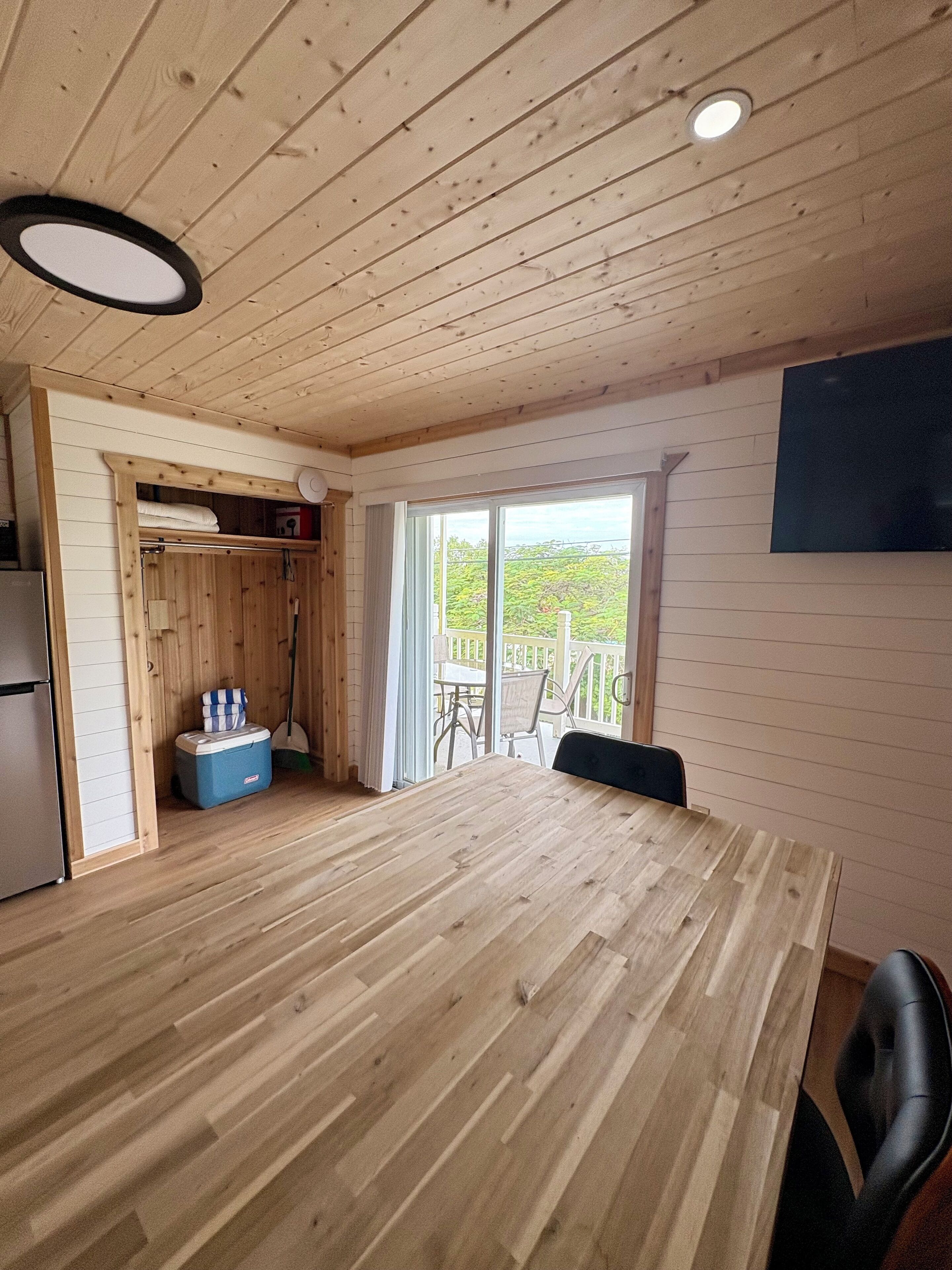 A spacious room with a wooden floor, a dining table, chairs, a refrigerator, a blue cooler, a ceiling light, and a large window with a view of greenery.