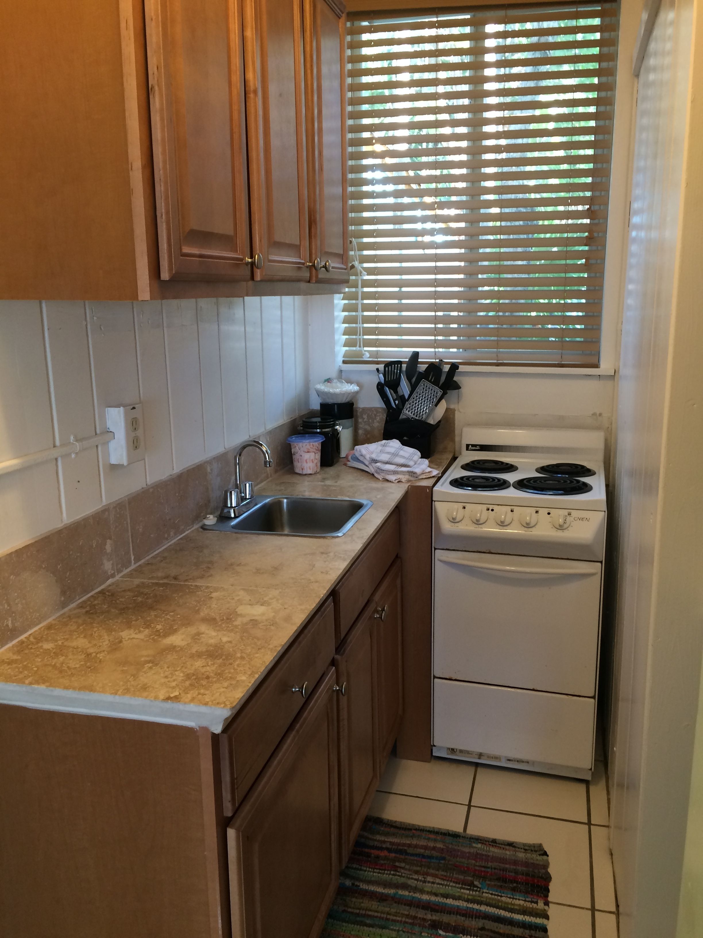 A small kitchen with wooden cabinets, a sink, a stove, and a window with blinds.