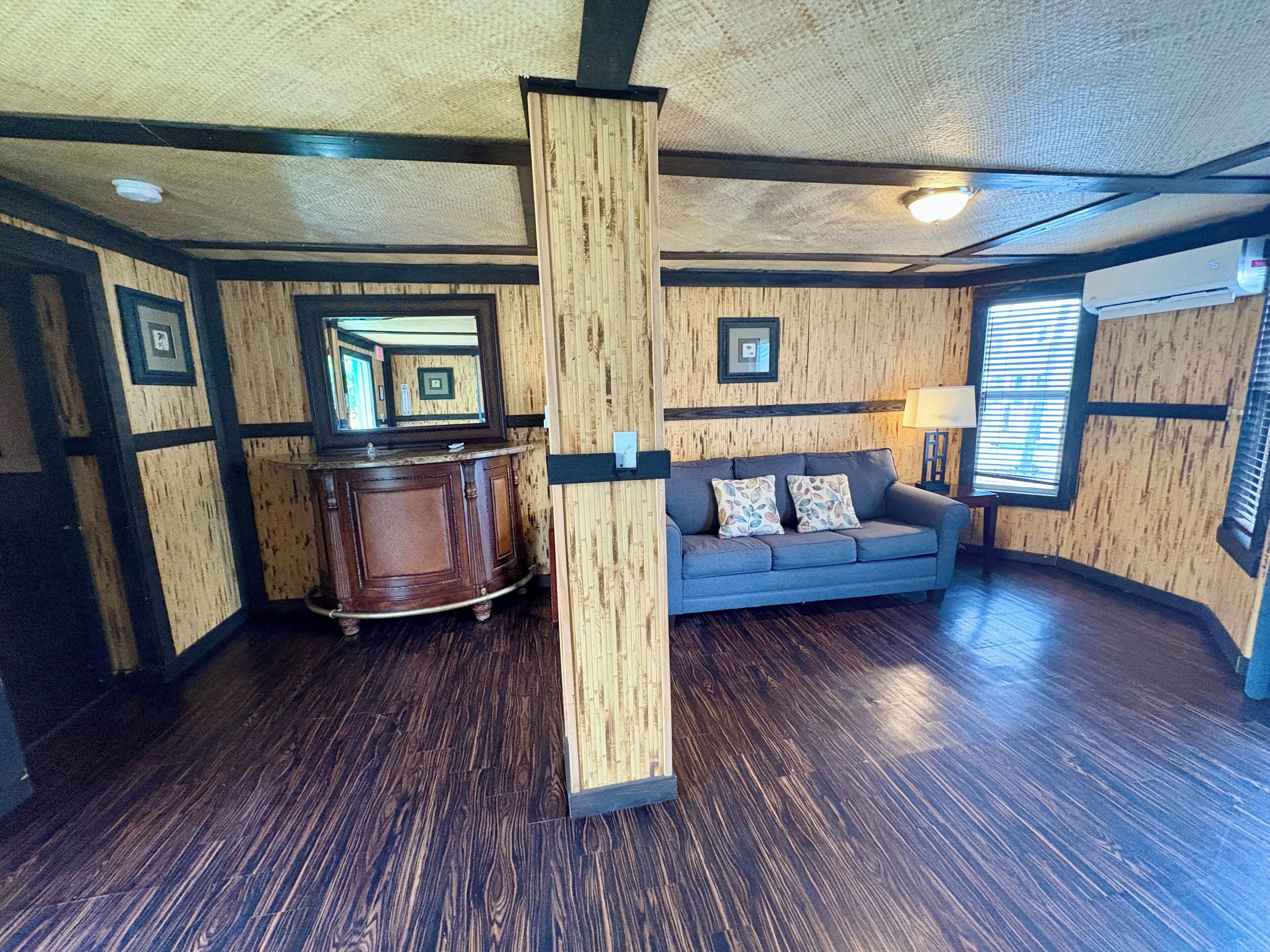 Una habitación con suelo de madera oscura, un sofá gris con cojines estampados, un pilar de madera y una pared de madera con cuadros enmarcados.