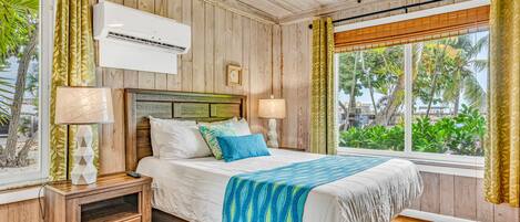 Un dormitorio con techo de madera, una cama con manta azul y blanca a rayas, dos lámparas de noche, una mesita de noche con cajones y una ventana con cortinas.