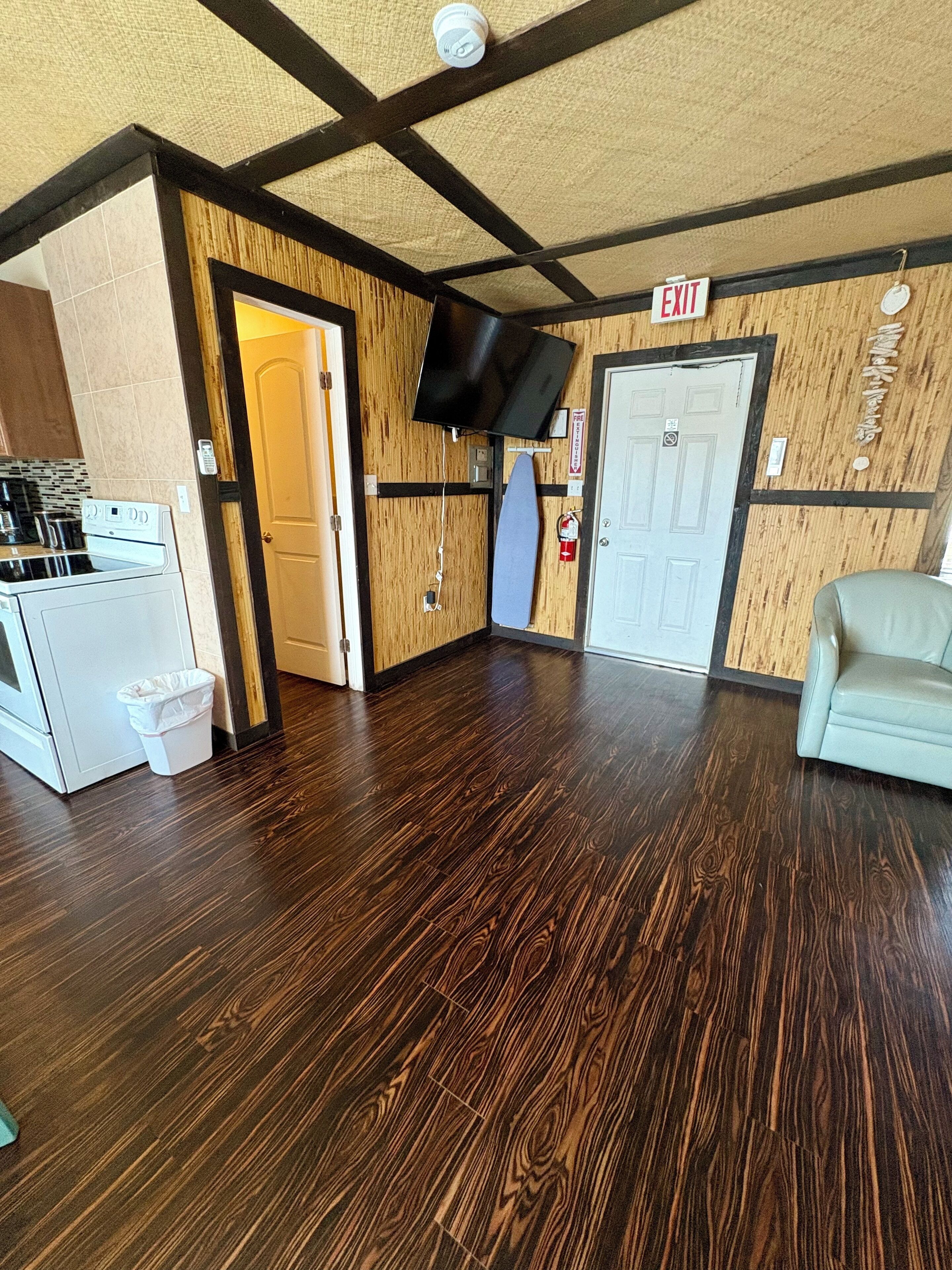 A room with a wooden floor, a flat-screen TV mounted on the wall, an exit sign, a door, and a small seating area with a chair.