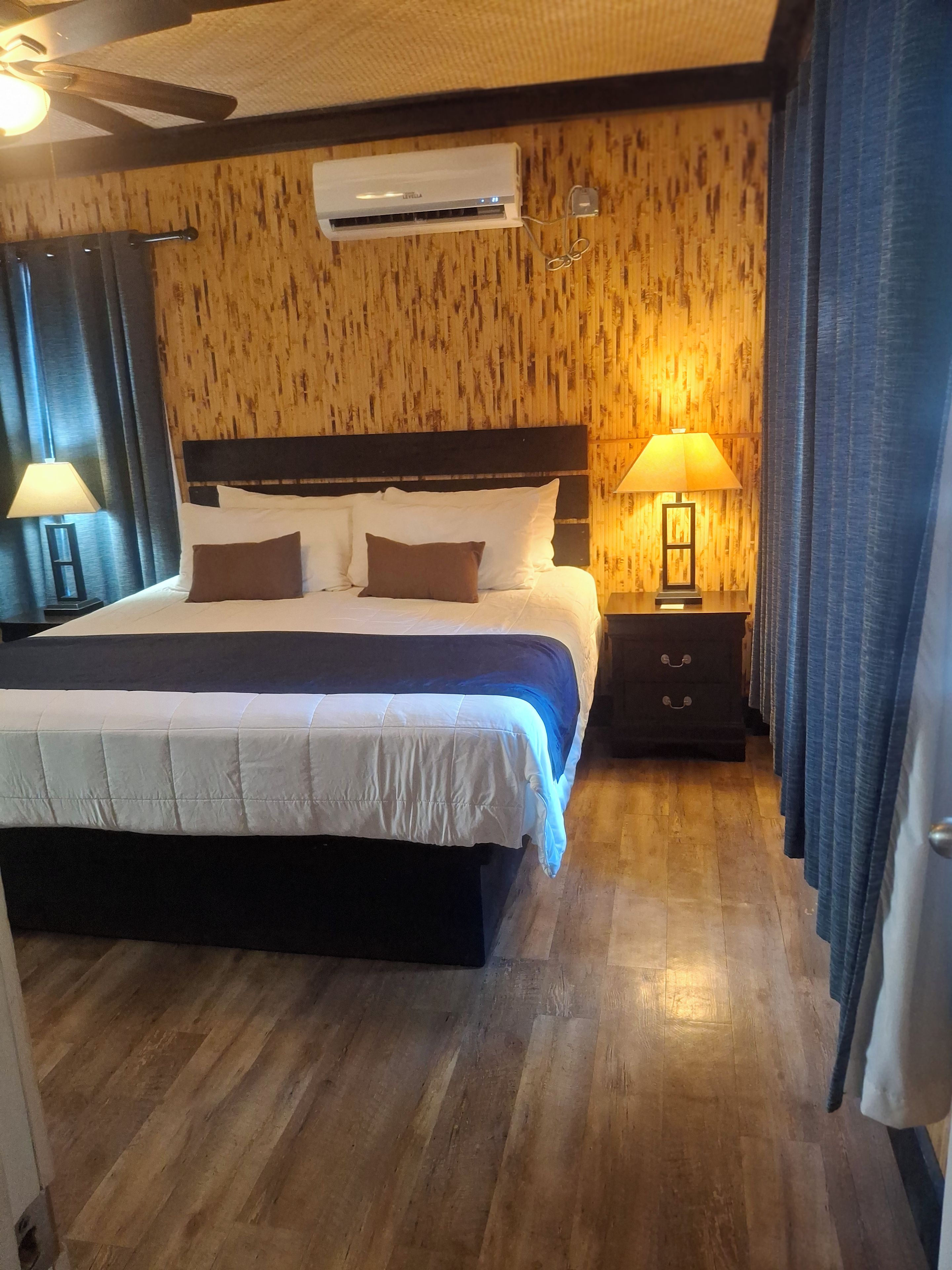 A hotel room with a large bed, wooden panel walls, an air conditioning unit, a nightstand with a lamp, and a wooden floor.