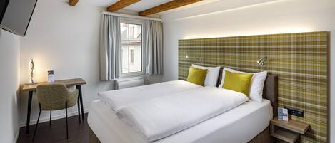 A hotel room with a large bed, a desk with a chair, a window with curtains, and a headboard with a plaid pattern.