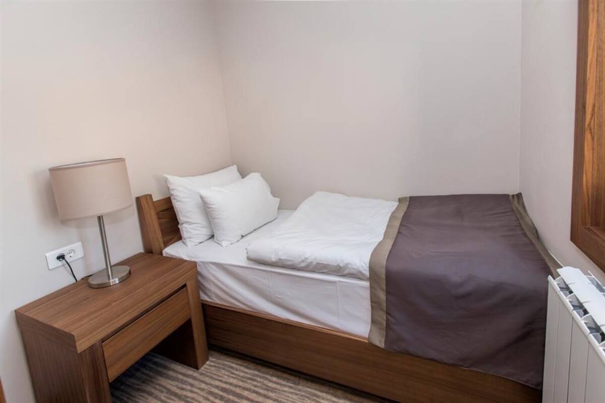 A single bed with white bedding and a brown throw, a bedside table with a lamp, and a power outlet on the wall.