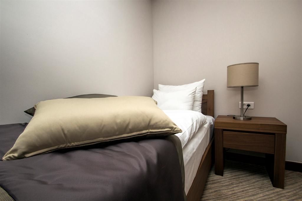 A neatly made bed with a beige pillow and a dark-colored bedspread in a room with a wooden nightstand and a lamp.