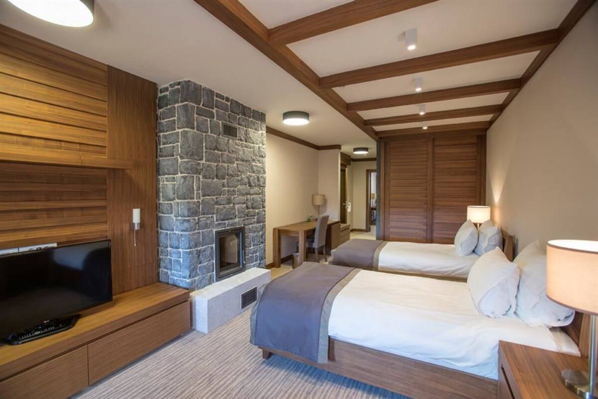 A hotel room with a stone fireplace, wooden beams, a TV, two beds, and a desk with a lamp.