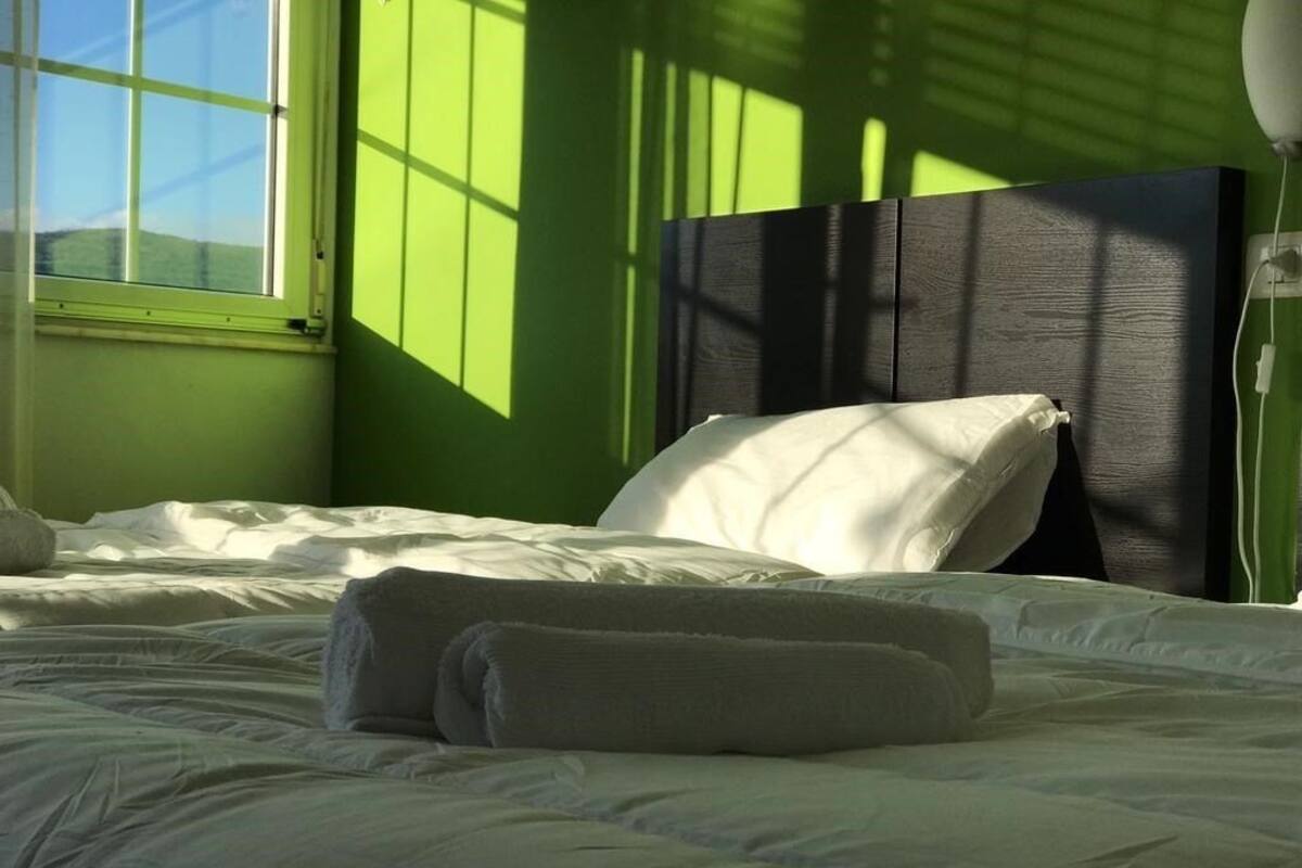 Ein ordentlich gemachtes Bett mit weißen Leinen und Kissen, eine Nachttischlampe und ein Fenster, durch das Sonnenschein Schatten an die Wand wirft.