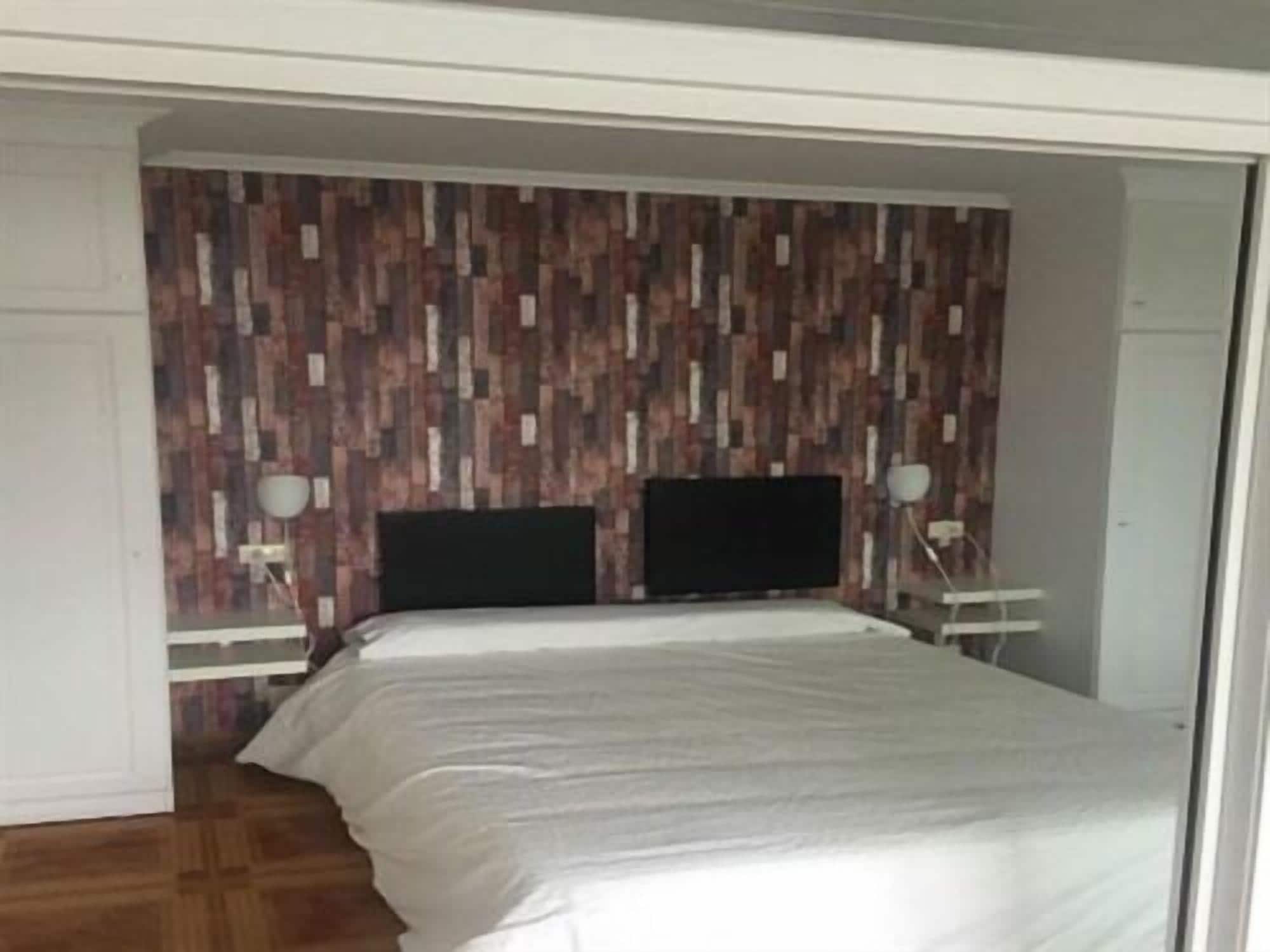 A bedroom with a wooden-patterned headboard, a bed with white bedding, two bedside tables with lamps, and a television on the headboard.