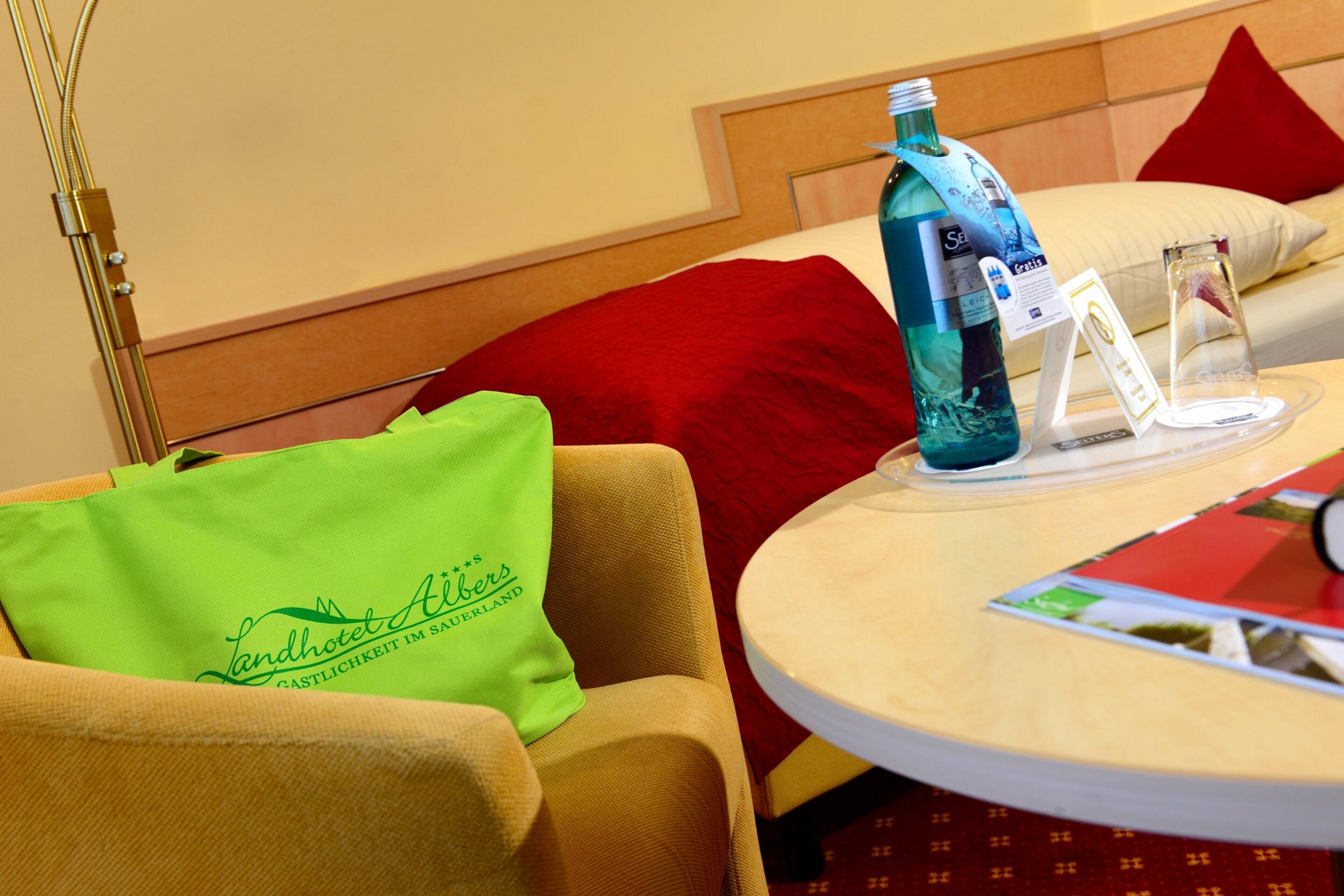 Habitación de hotel con cama, una mesa redonda con una botella y un vaso, y un almohadón verde con texto.