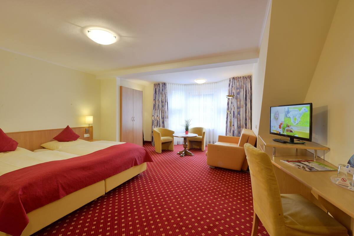 A hotel room with a large bed, a desk with a television, a small table with chairs, and a window with curtains.