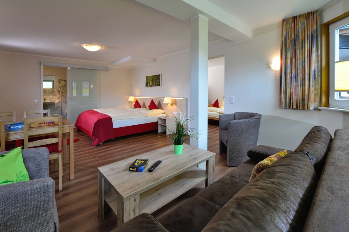 A hotel room with a bed, a sofa, a coffee table, a chair, and a small table with a remote control.