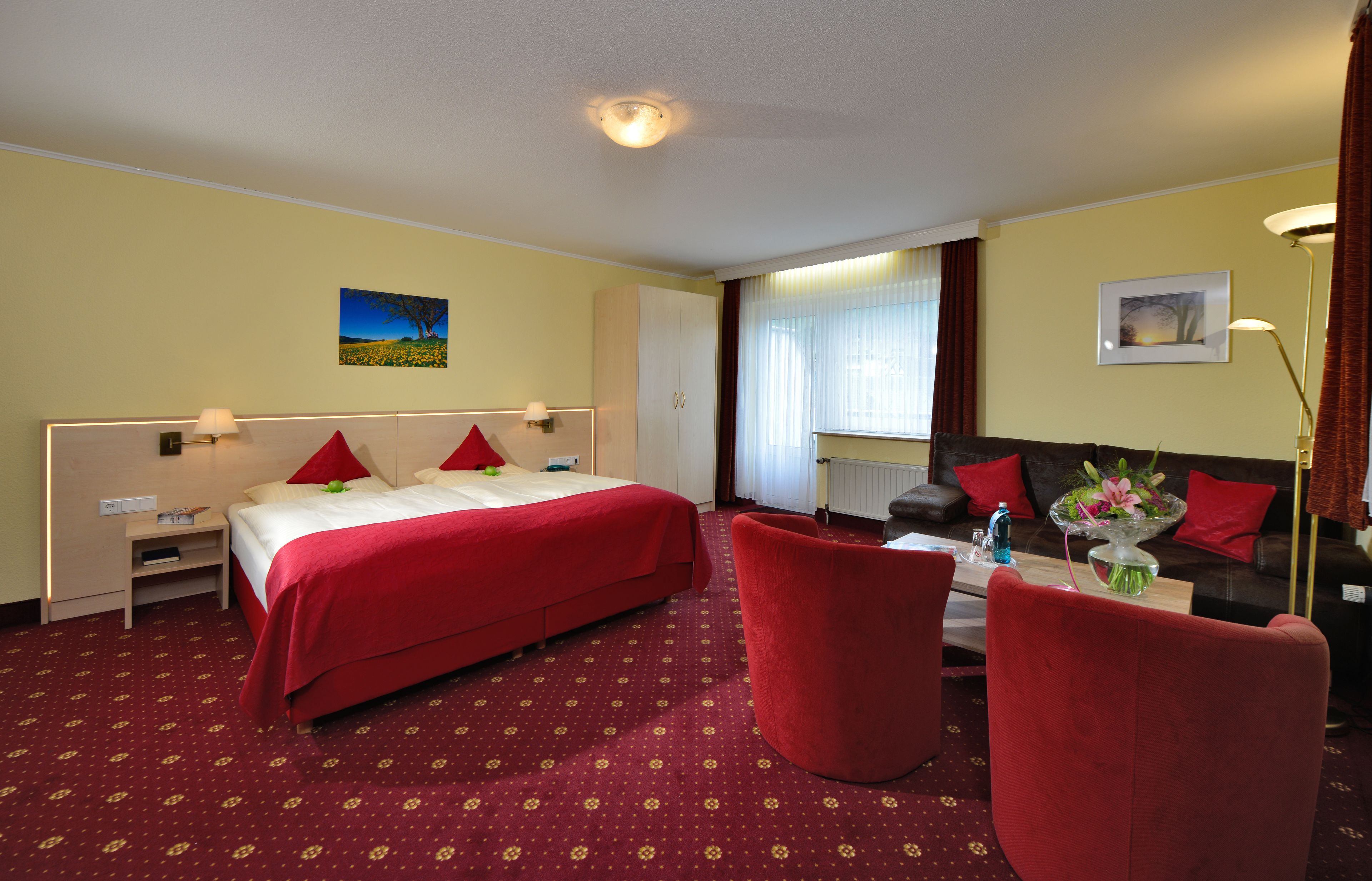 A hotel room with a bed, two red chairs, a sofa, a table with a vase, and a painting on the wall.