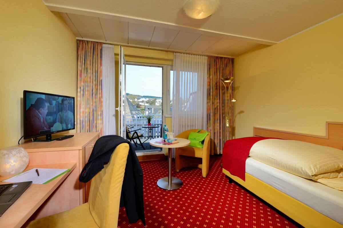 A hotel room with a bed, a desk with a laptop, a television, a chair, and a small table.