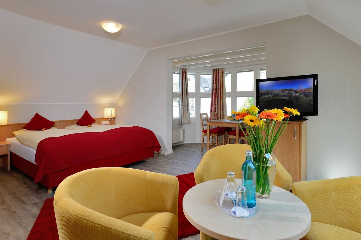 A hotel room with a bed, two yellow armchairs, a small table with a vase of flowers, a television, and a window with curtains.