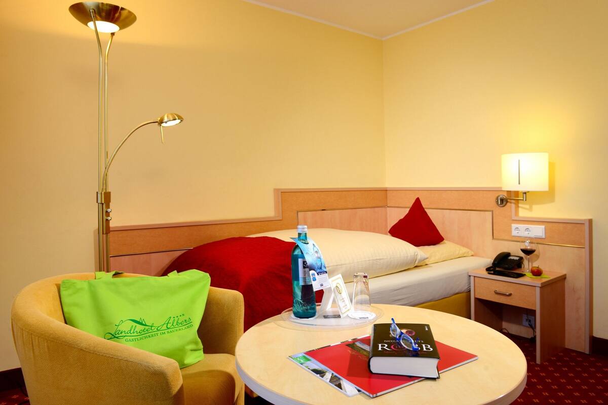 A hotel room with a bed, bedside table, lamp, chair, and a round table with a book and a bottle.