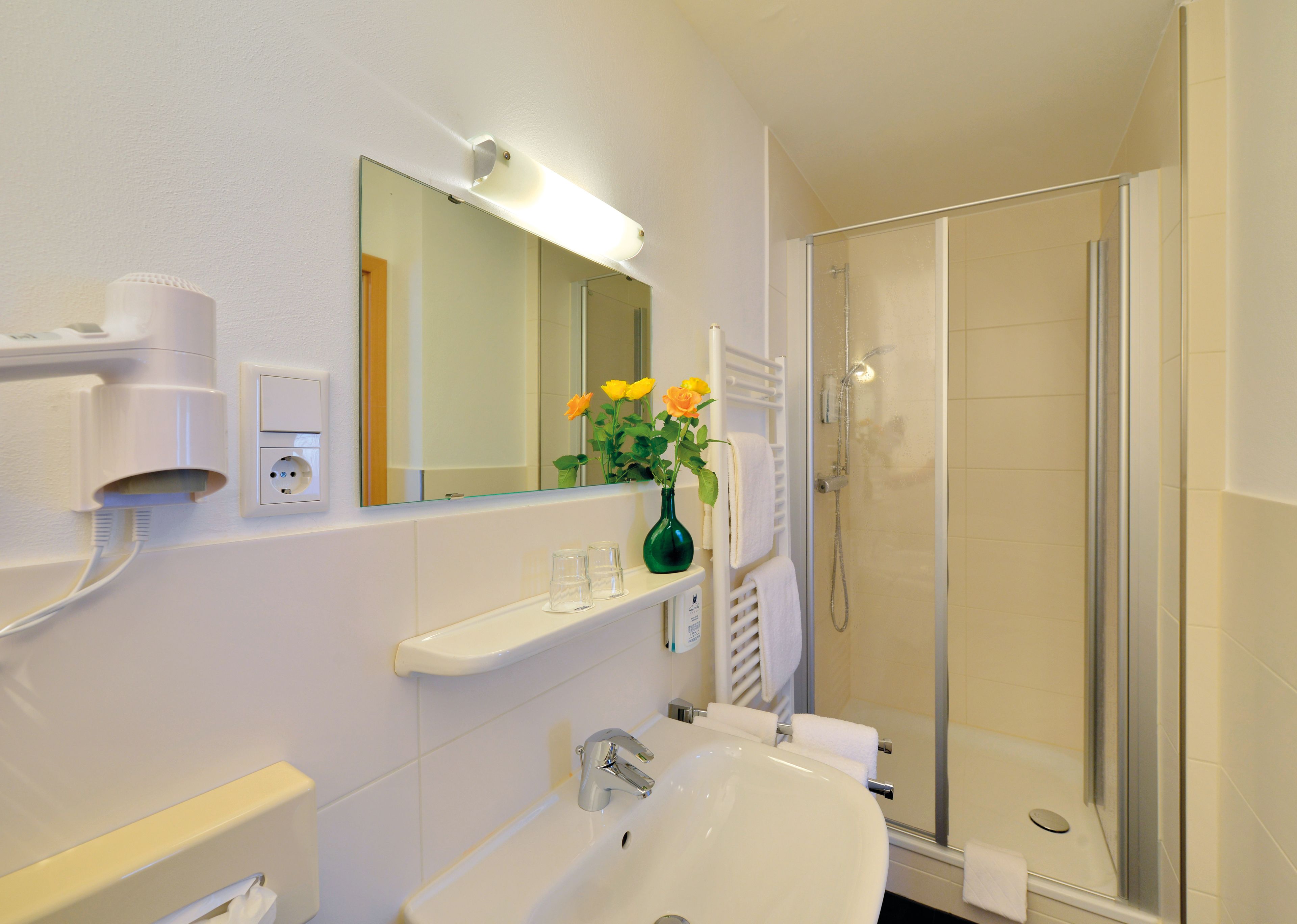 Un baño con lavamanos, espejo y ducha. Hay un florero con flores sobre el lavamanos y un perchero encima del espejo.