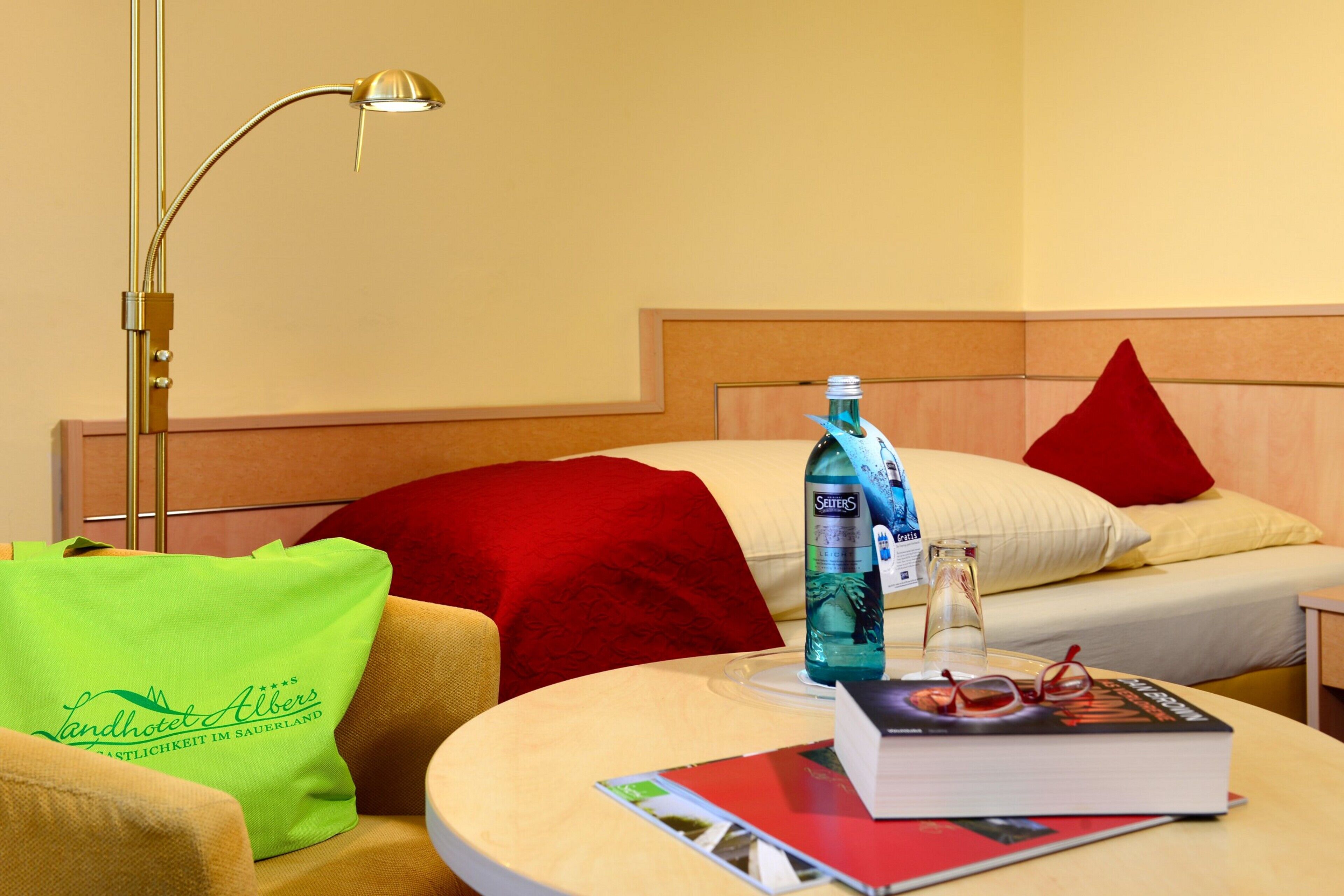 A hotel room with a bed, a bedside table with a book and glasses, a bottle, a lamp, and a green bag.